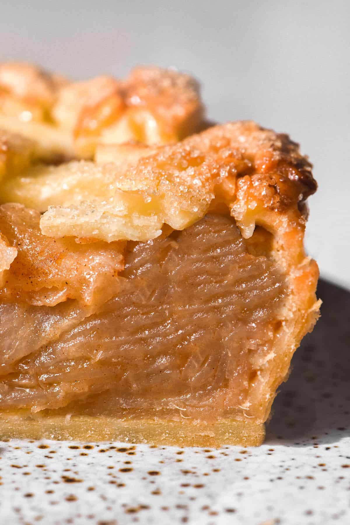 A macro side on image of a slice of low FODMAP apple pie. The crust is a deep golden brown and topped with raw sugar. The slice sits on a white speckled ceramic plate against a white backdrop. 