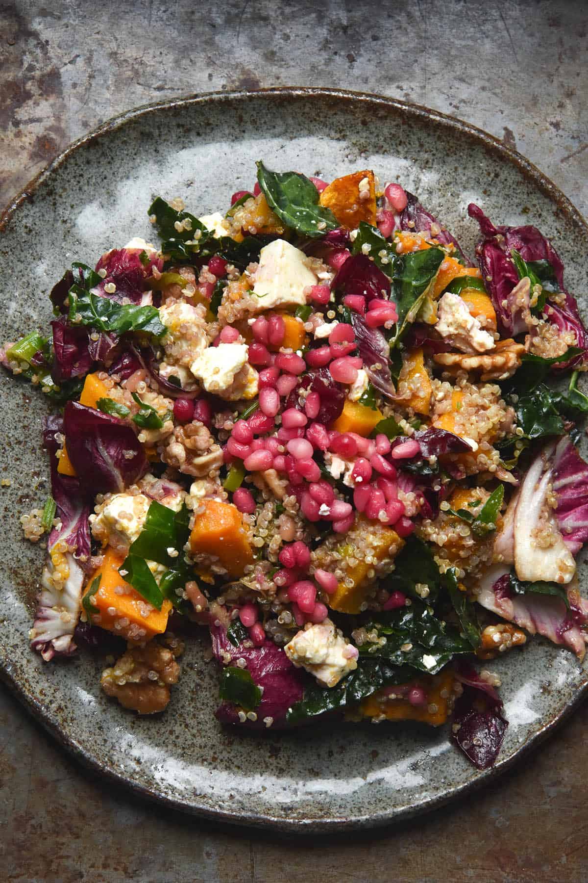 An aerial image of a dark blue ceramic plate topped with a roast pumpkin, quinoa, feta and kale salad topped with pomegranates and walnuts. The salad sits on a dark grey mottled steel backdrop.