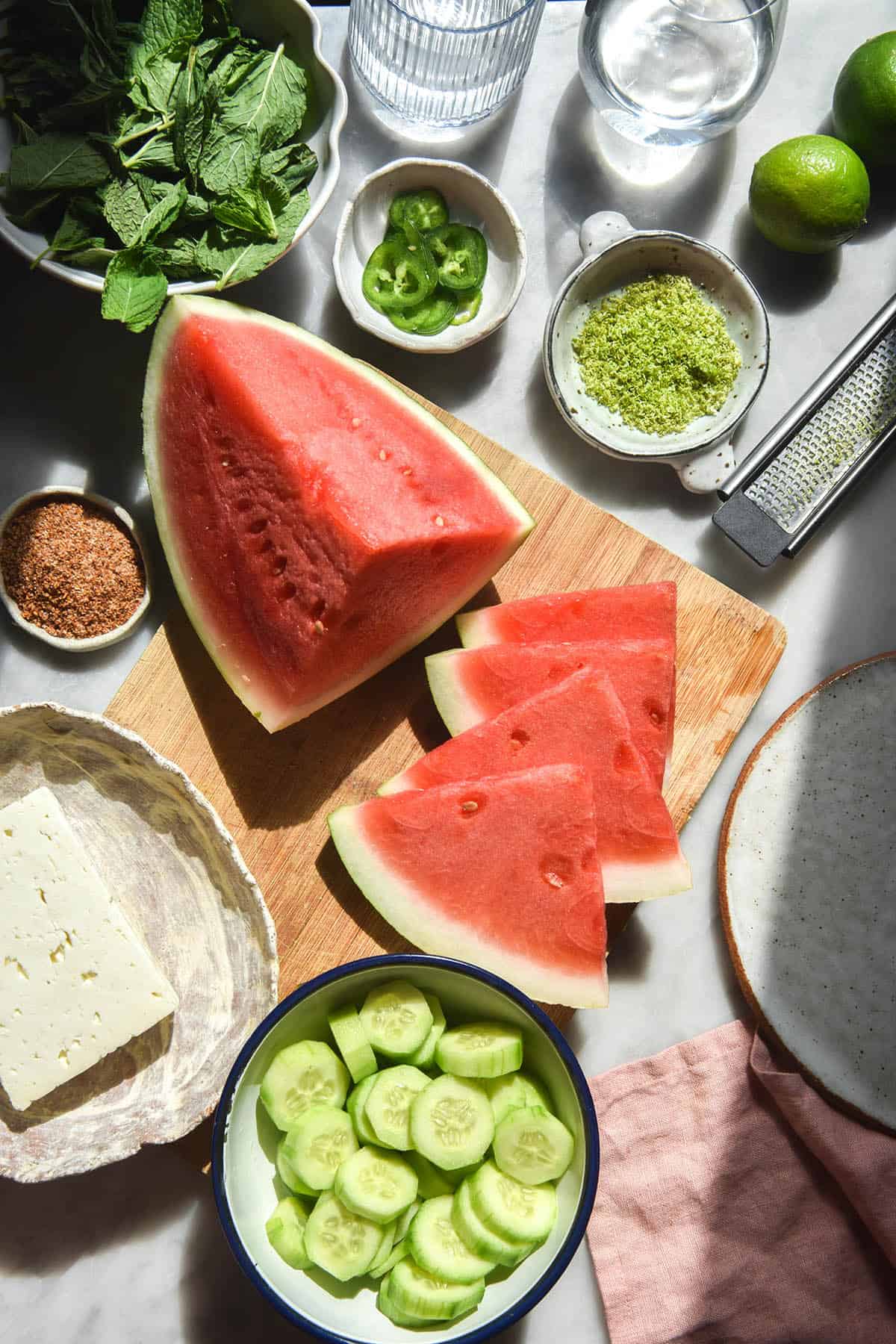 An aerial sunlit image of the ingredients for a watermelon and tajin salad casually arranged on a white marble table. The sliced watermelon sits in the centre, surrounded by cucumber, feta, tajin, mint, jalapeno slices and lime zest.