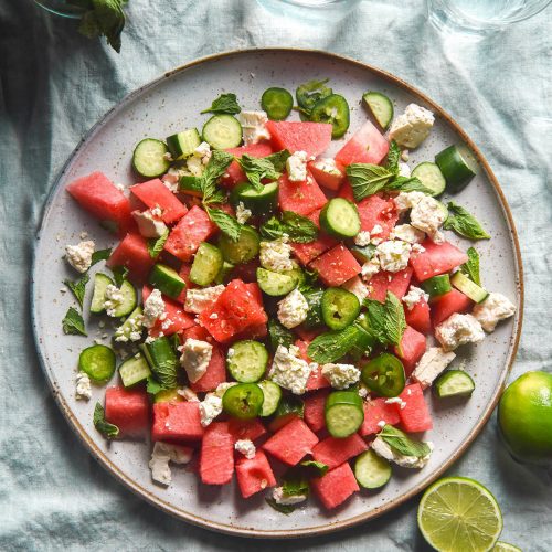 An aerial image of a plate of watermelon tajin salad on a light green linen tablecloth. The salad is surrounded by glasses of water, extra mint and limes.