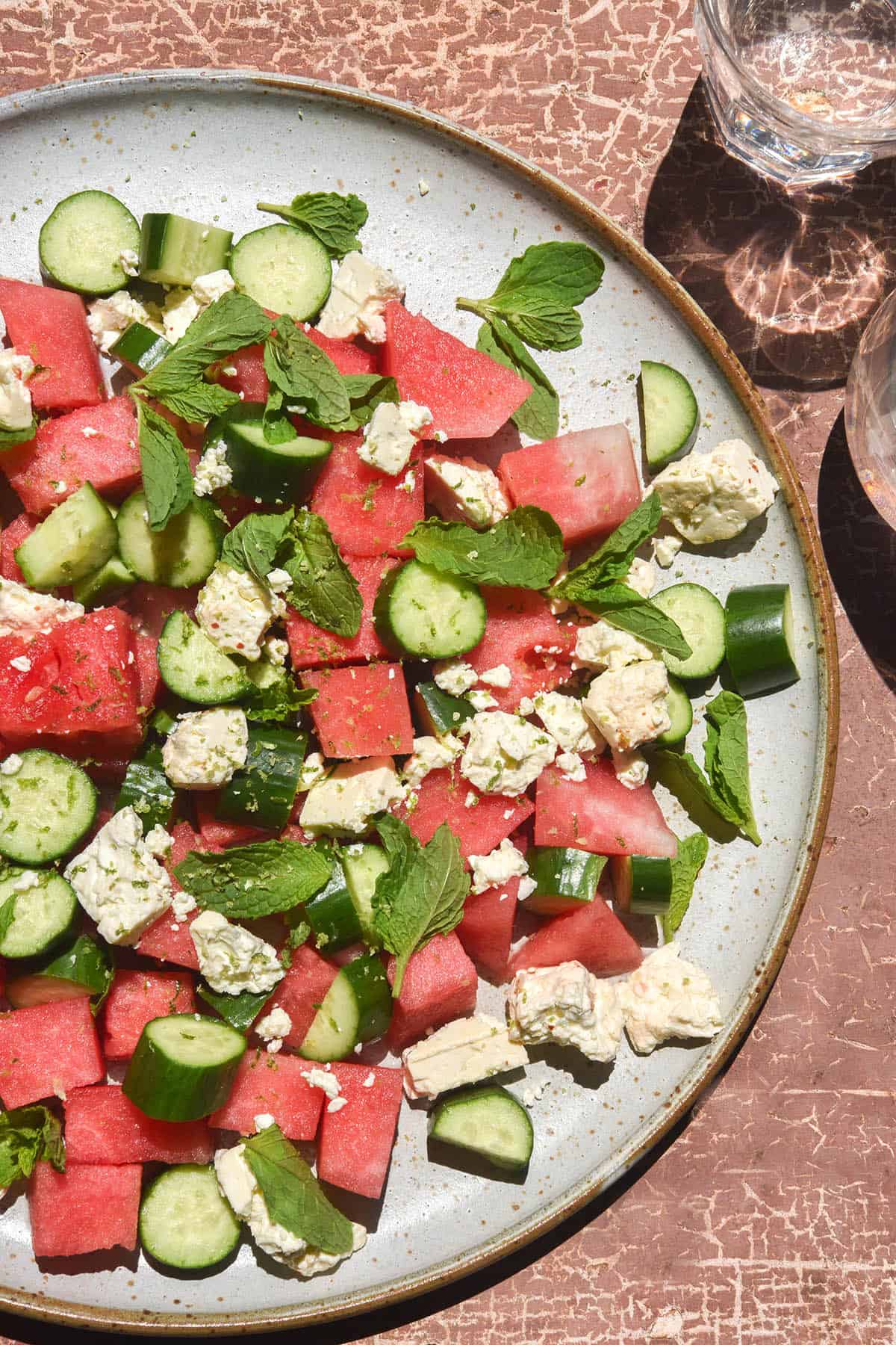 A brightly lit aerial image of a watermelon salad with tajin, cucumber, feta and mint atop a white ceramic serving plate on a rustic rose coloured backdrop