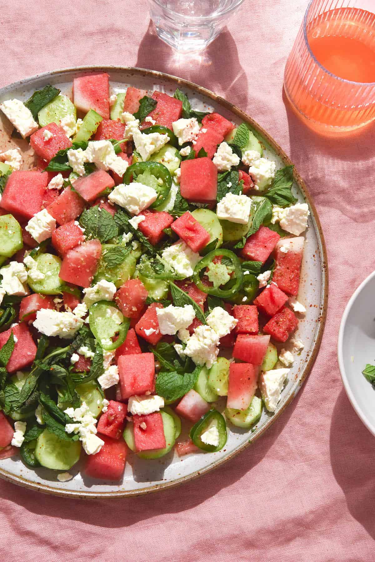 A brightly lit aerial image of a watermelon salad with tajin, cucumber, mint, feta and jalapenos. The salad sits on a white ceramic plate atop a pale pink linen backdrop.