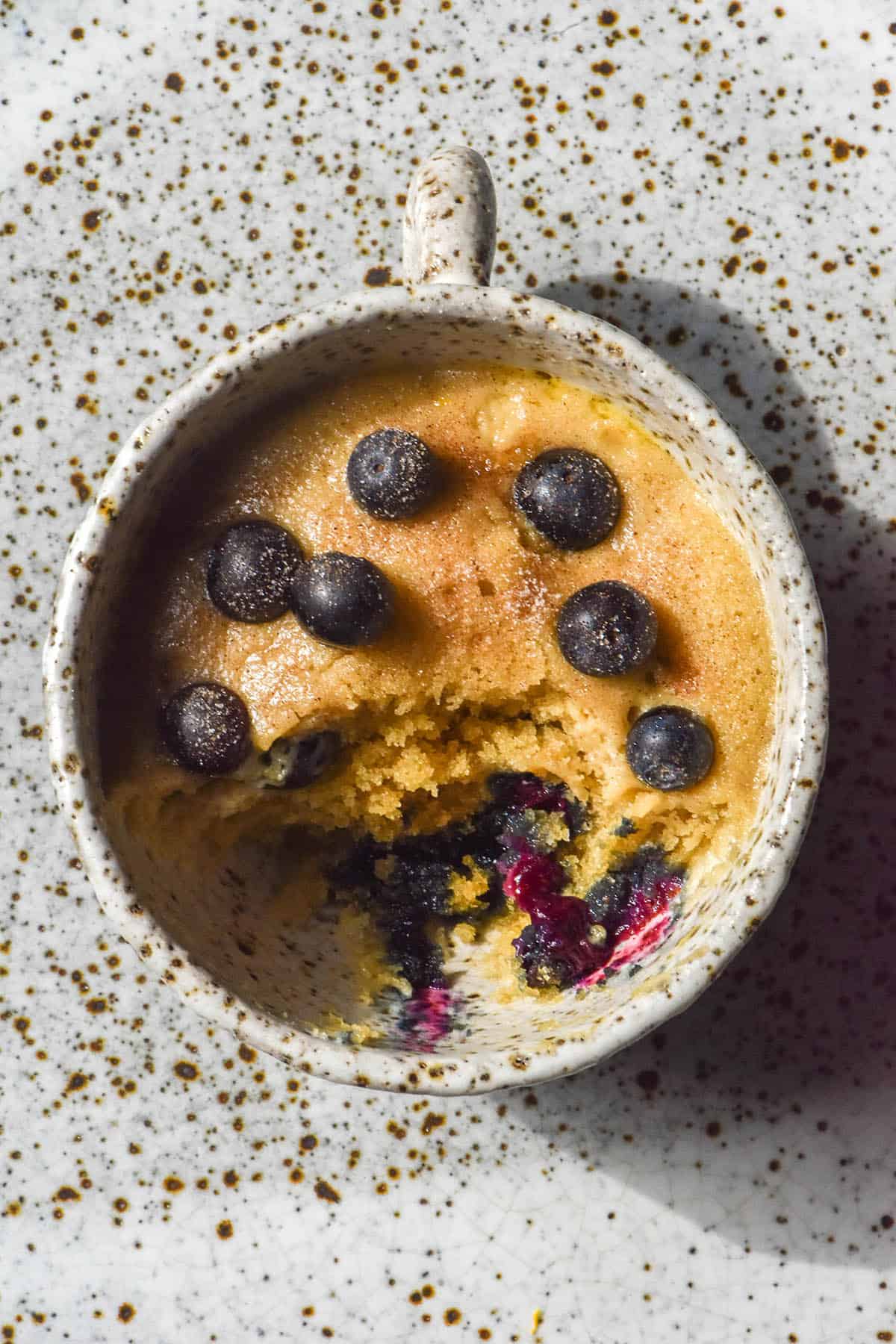 An aerial image of a gluten free blueberry mug cake on a white speckled ceramic plate