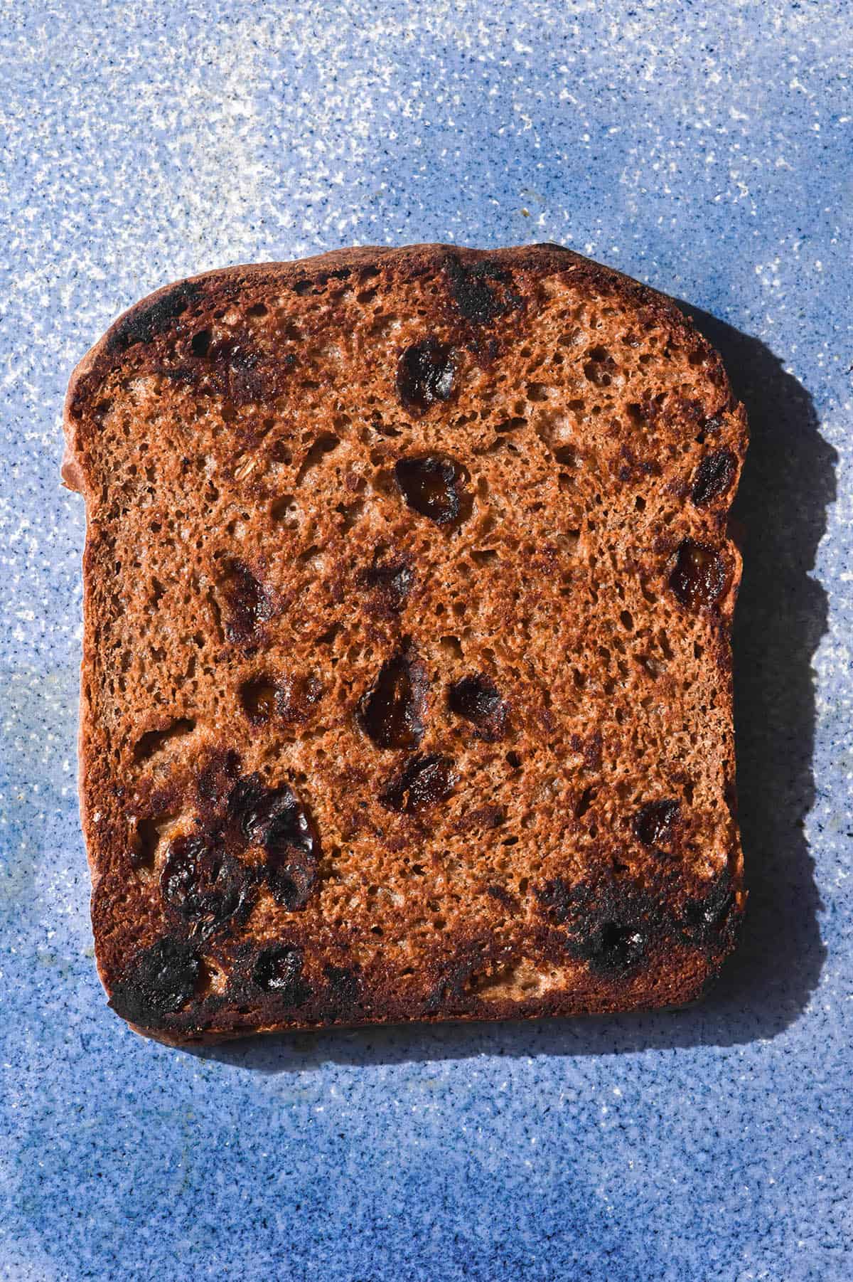 A brightly lit aerial image of a slice of buckwheat cinnamon raisin loaf atop a bright blue ceramic plate. 