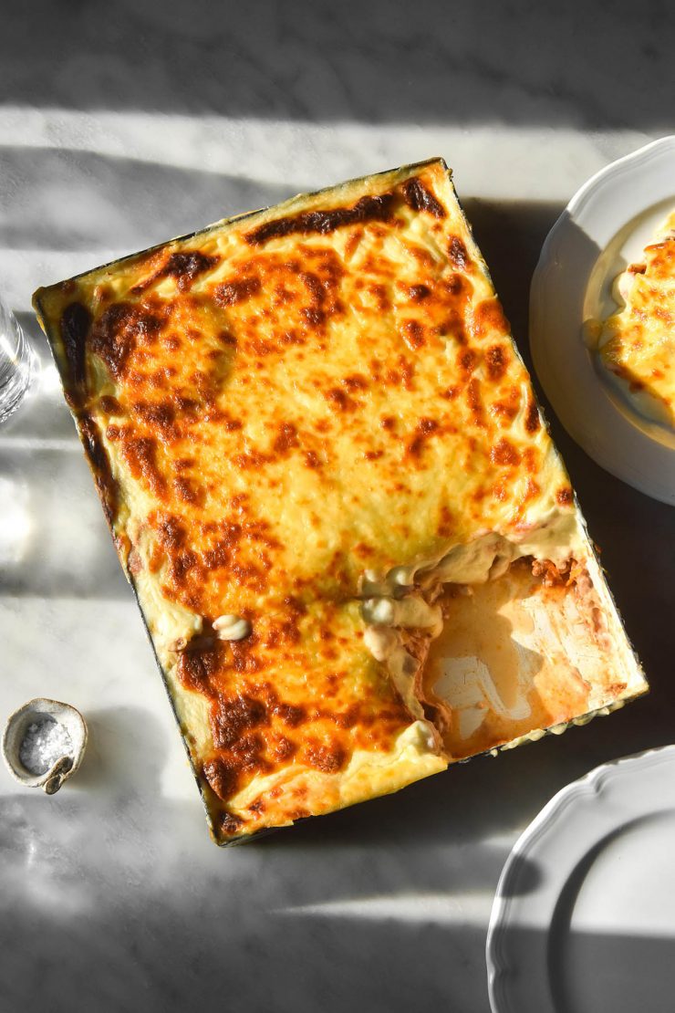 An aerial view of a low FODMAP lasagne on a white marble table in contrasting sunlight. The lasagne is surrounded by glasses of water and extra plates of lasagne