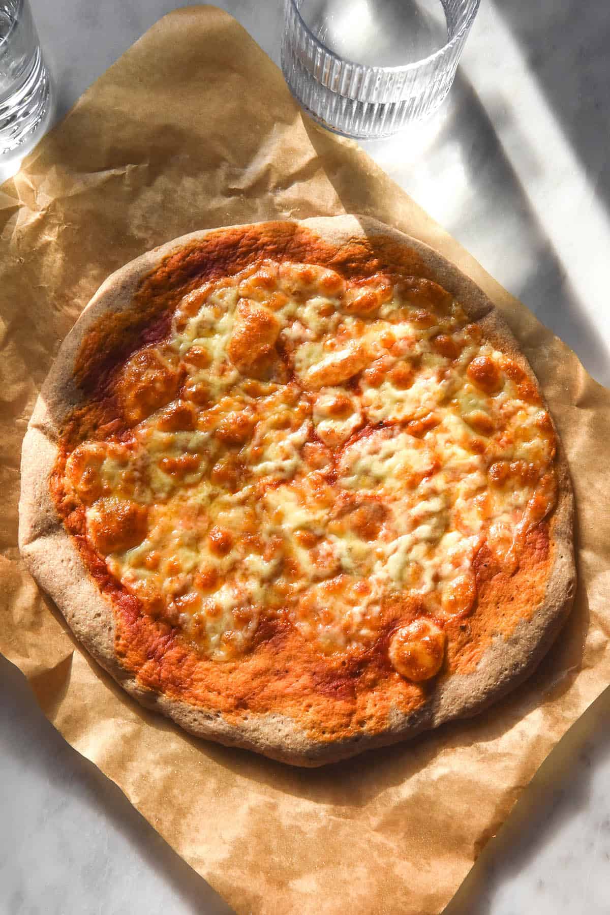 An aerial sunlit image of a buckwheat flour pizza on a sheet of baking paper atop a white marble table. The pizza is topped with margherita toppings