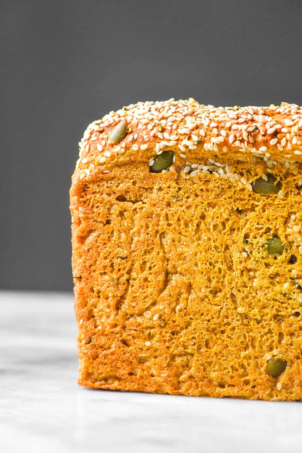 A side on view of a loaf of gluten free pumpkin bread with seeds on a white marble table against a white backdrop