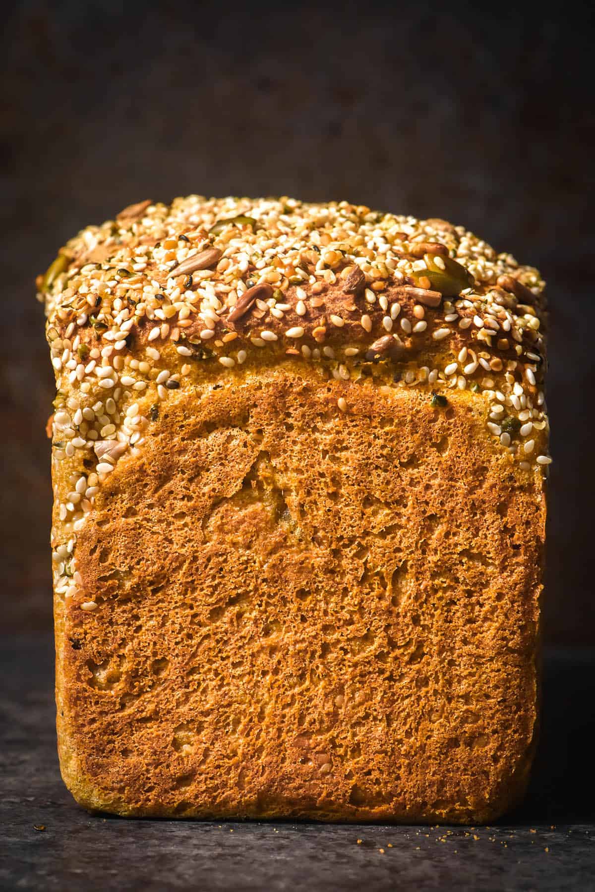 A side on image of a loaf of savoury buckwheat pumpkin bread topped with mixed seeds. The bread sits atop a dark tray against a dark backdrop. 