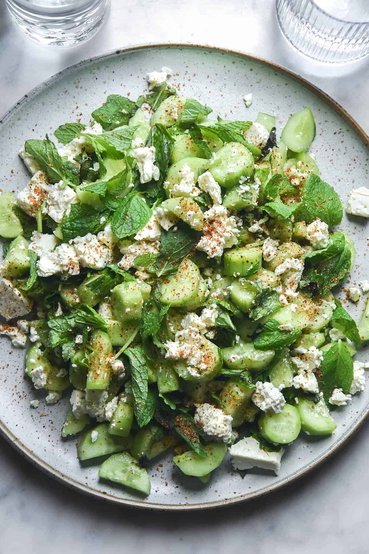 A sunlit aerial image of a cucumber, tajin, feta and mint salad on a white speckled ceramic plate atop a white marble table. 