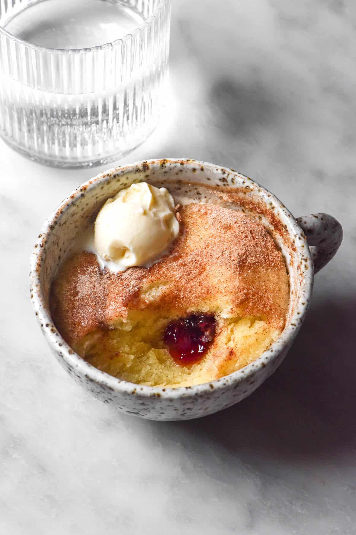 A side on image of a gluten free jam donut mug cake in a white speckled ceramic mug atop a white marble table