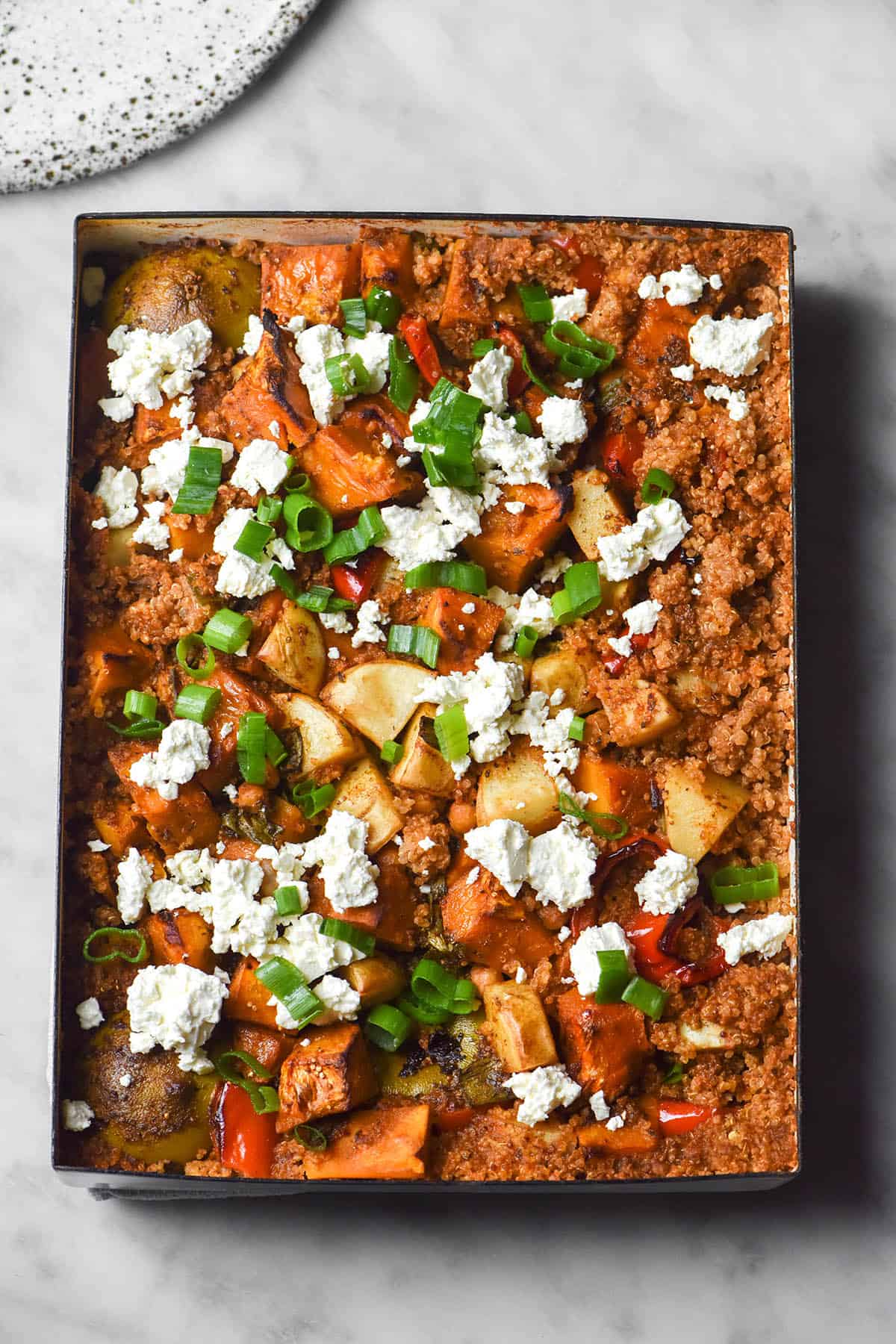 An aerial image of a low FODMAP quinoa tray bake with vegetables, chickpeas, Mexican spices and limes, topped with feta and spring onion greens. The bake sits on a white marble table and a white speckled ceramic plate sits to the top left of the image.