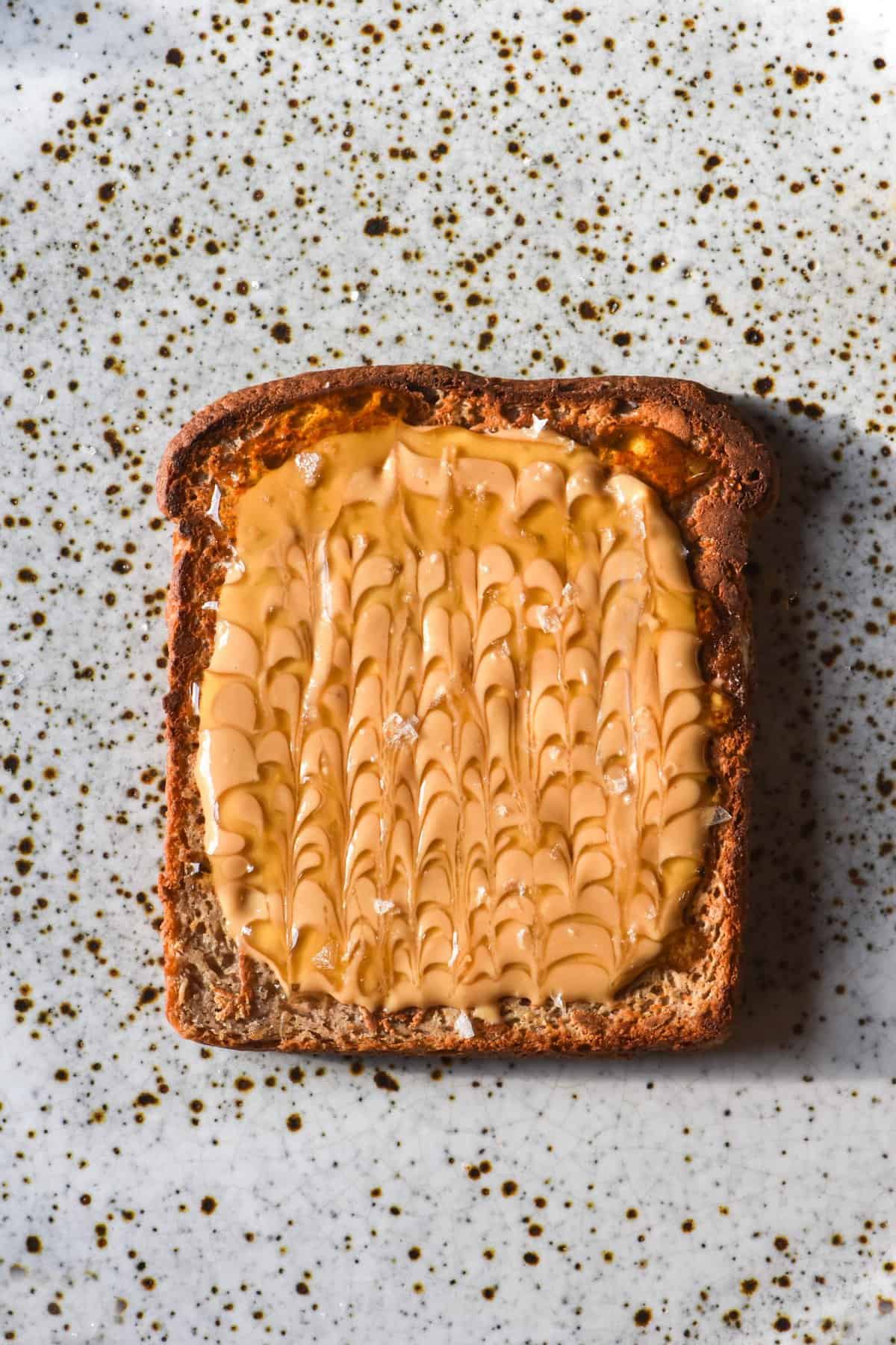 An aerial image of a slice of gluten free vegan high protein bread toasted and topped with peanut butter and rice malt syrup. The toast sits on a white speckled ceramic plate.