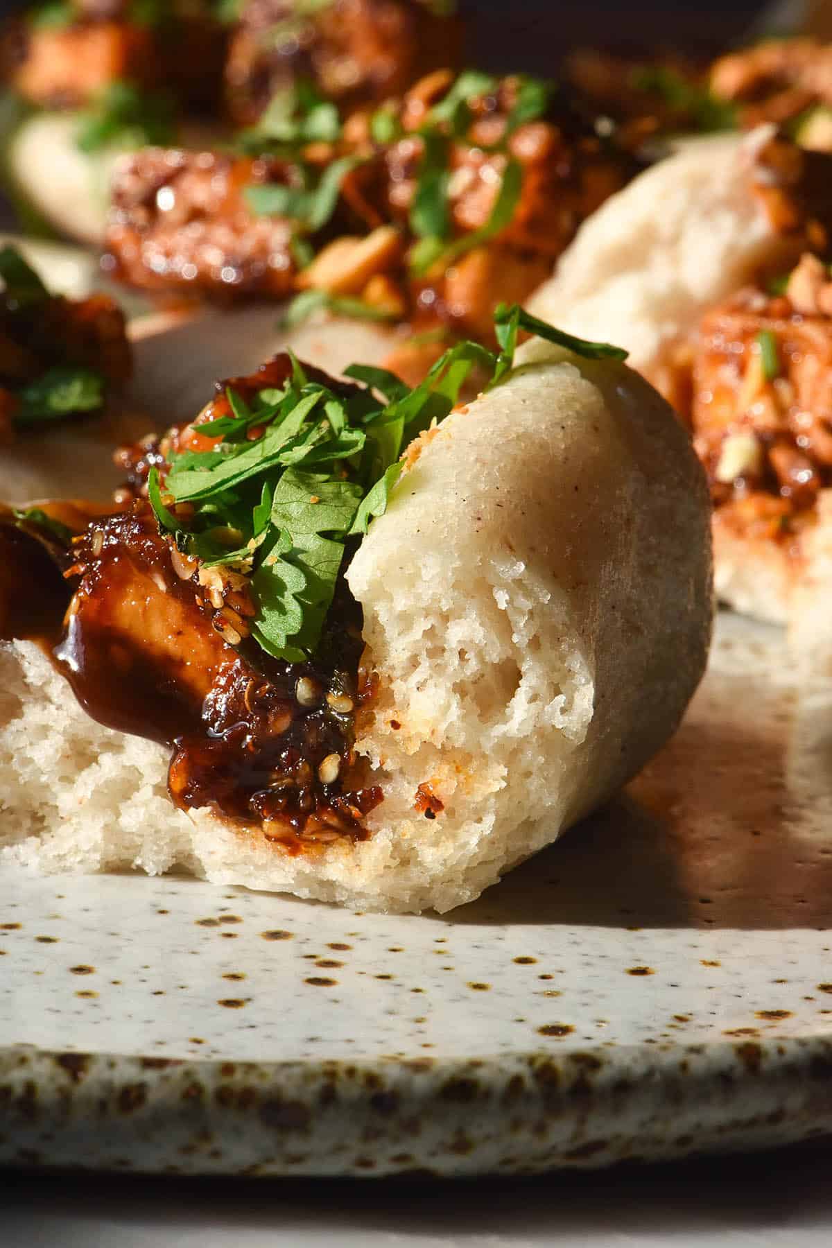 A close up image of gluten free bao buns on a white speckled ceramic plate. The front bao bun has been bitten into, revealing the soft white crumb and the saucy tofu filling. 