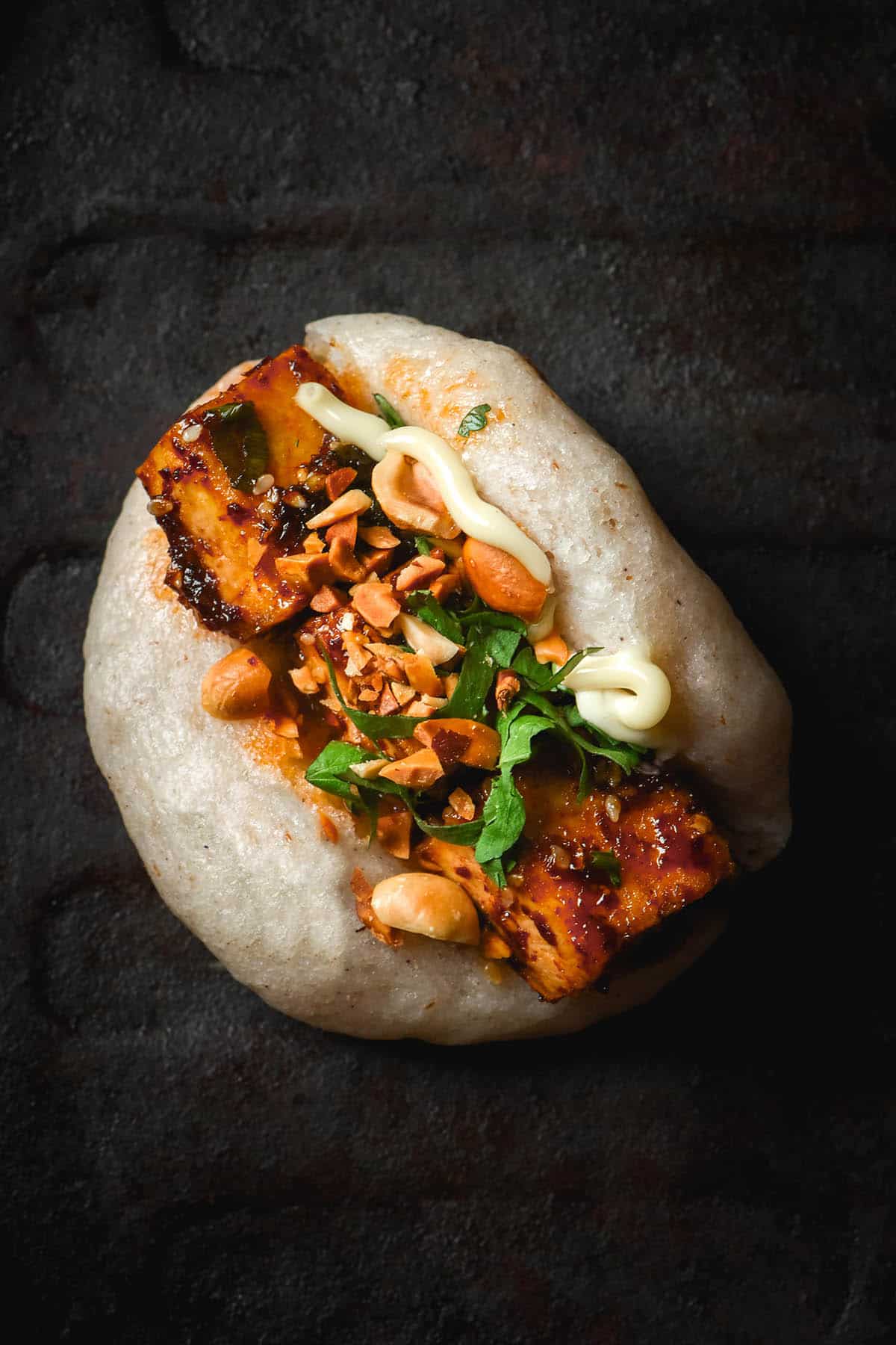 An aerial image of a gluten free bao bun on a dark steel plate. The bao bun is topped with chilli tofu, coriander, peanuts and a squeeze of kewpie mayonnaise