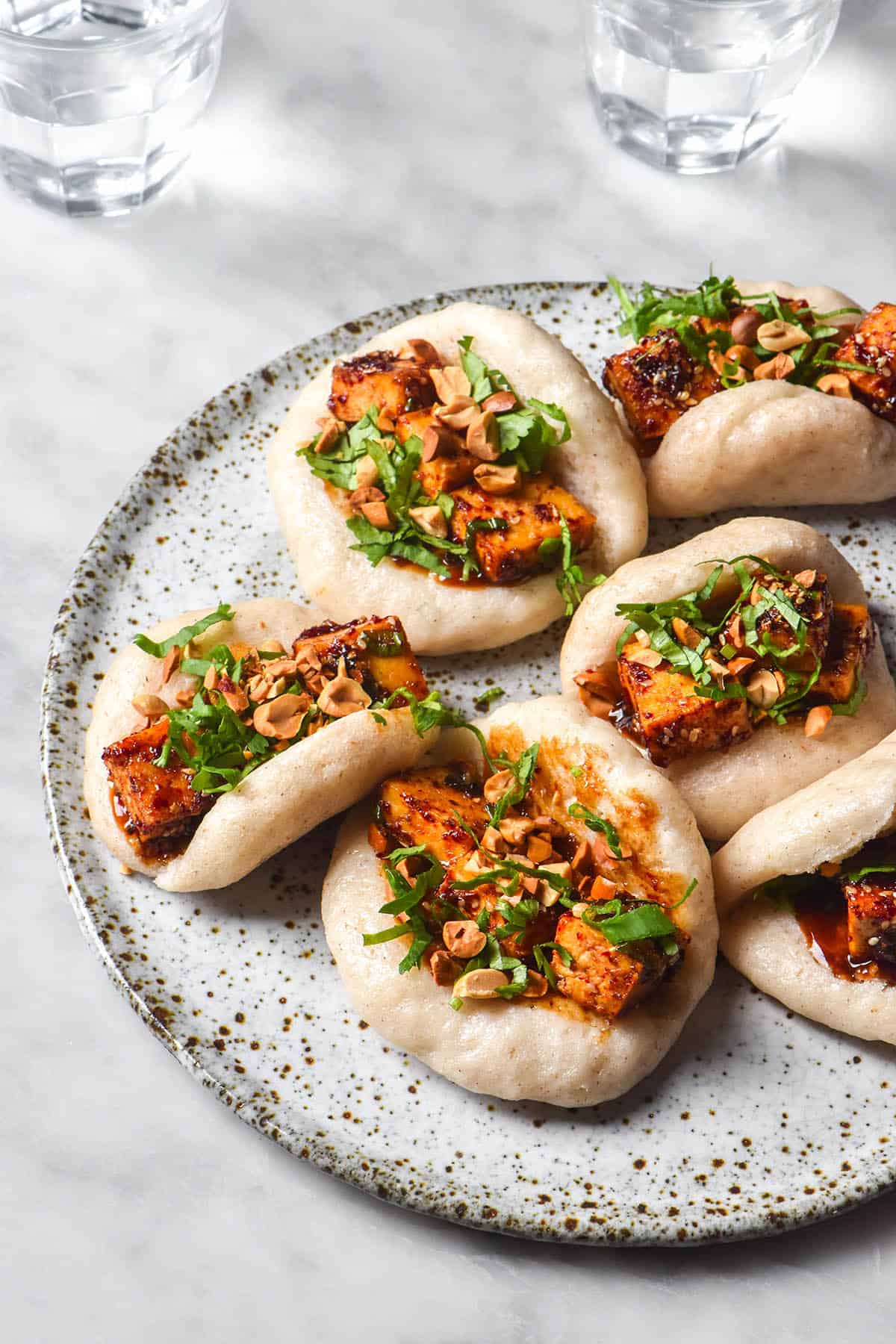 An aerial side on image of a plate of gluten free bao buns topped with chilli oil tofu, hoisin sauce, chopped coriander and peanuts. The buns sit atop a white ceramic plate on a white marble table. 
