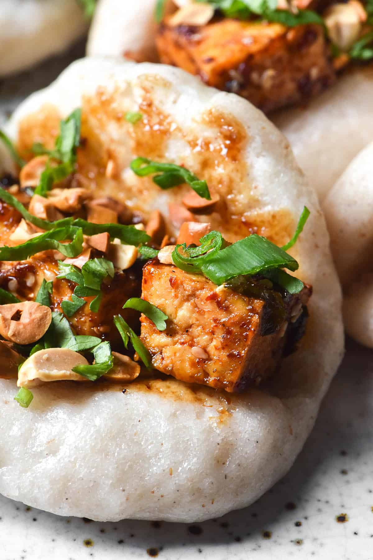 A macro close up of a gluten free bao bun filled with chilli tofu, hoisin sauce, peanuts and spring onion greens