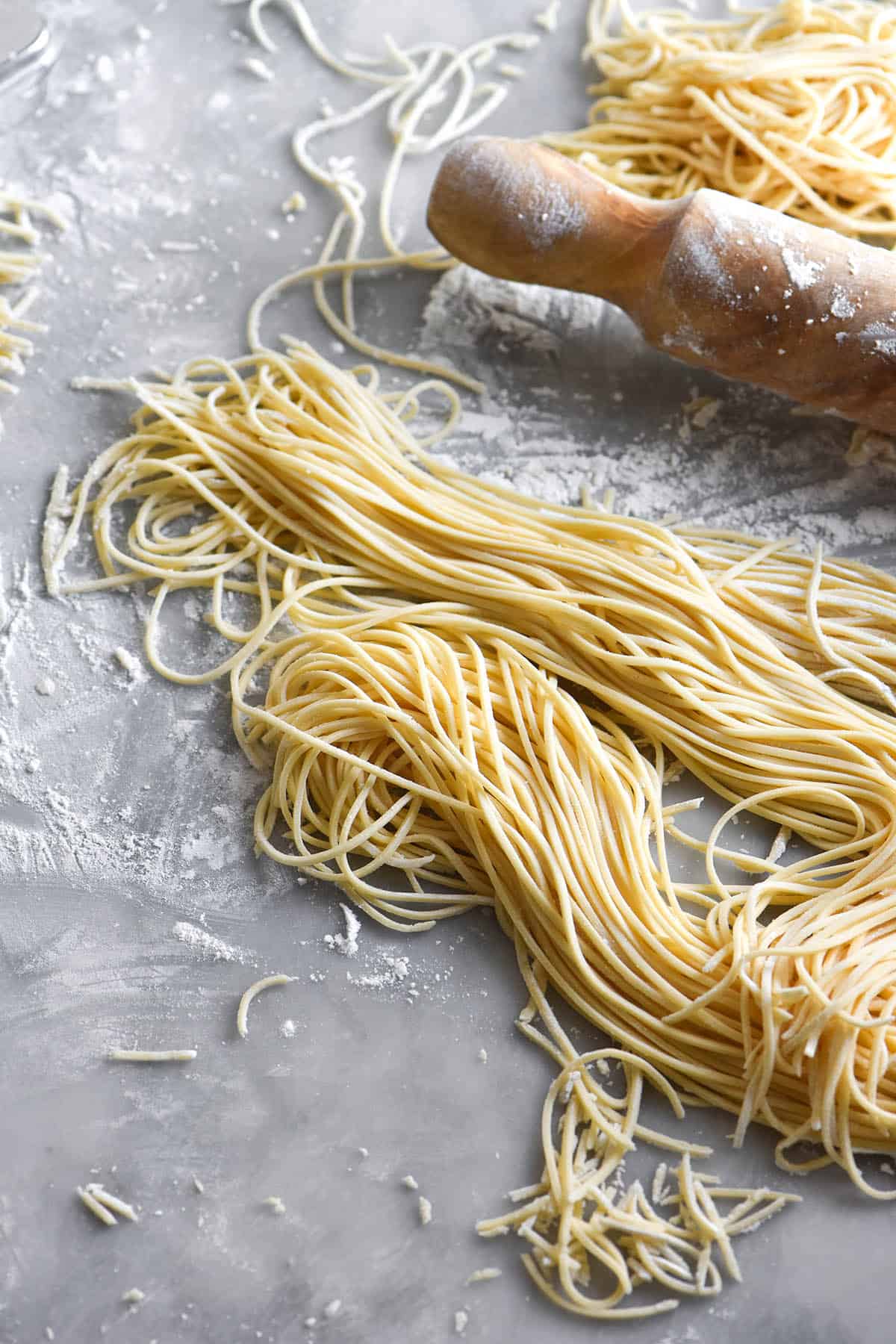 A messy image of gluten free egg noodles being made on a white marble table. They are casually arranged in flowing piles of noodles and a rolling pin sits to the back of the image