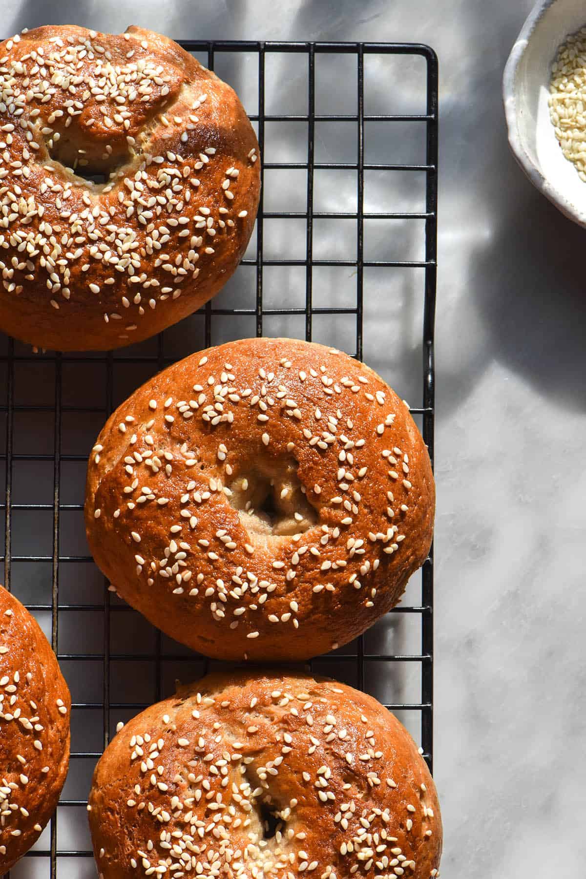 An aerial close up image of gluten free bagels topped with sesame seeds on a rooling rack atop a sunlit white marble table. A white bowl of sesame seeds sits to the top right of the image