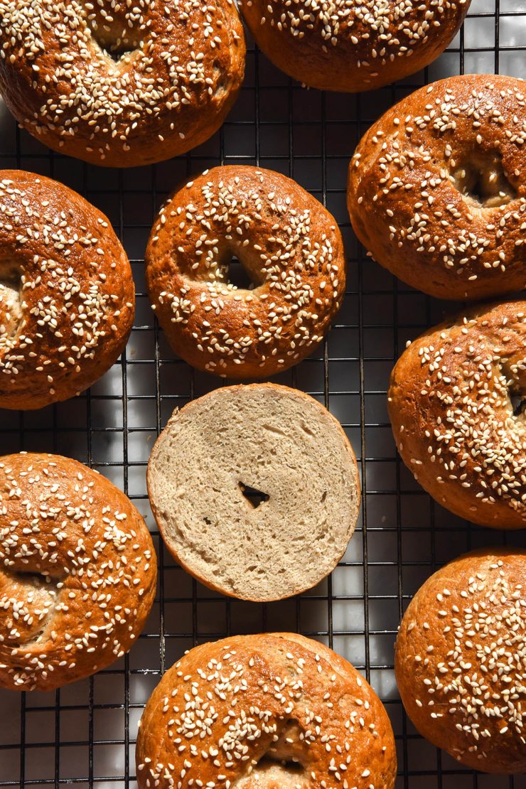 An aerial sunlit image of gluten free high protein bagels topped with sesame seeds sitting on a cooling rack atop a white marble table