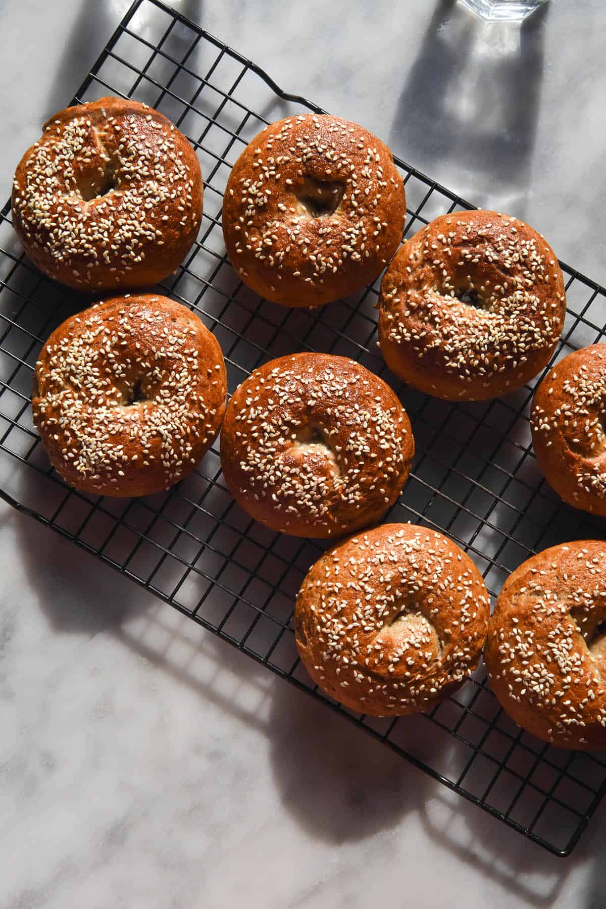 An aerial sunlit image of gluten free high protein bagels topped with sesame seeds sitting on a cooling rack atop a white marble table
