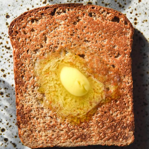 A brightly lit aerial image of a piece of low FODMAP high protein toast topped with melting butter. The toast sits atop a white speckled ceramic plate