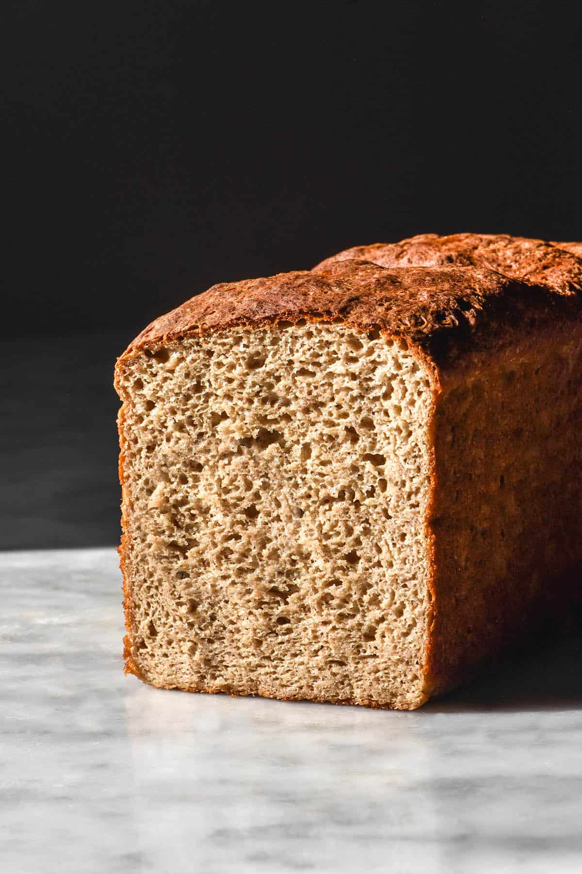 A side on image of a loaf of high protein buckwheat bread on a white marble table in contrasting sunlight. The loaf has been sliced to reveal the inner crumb.