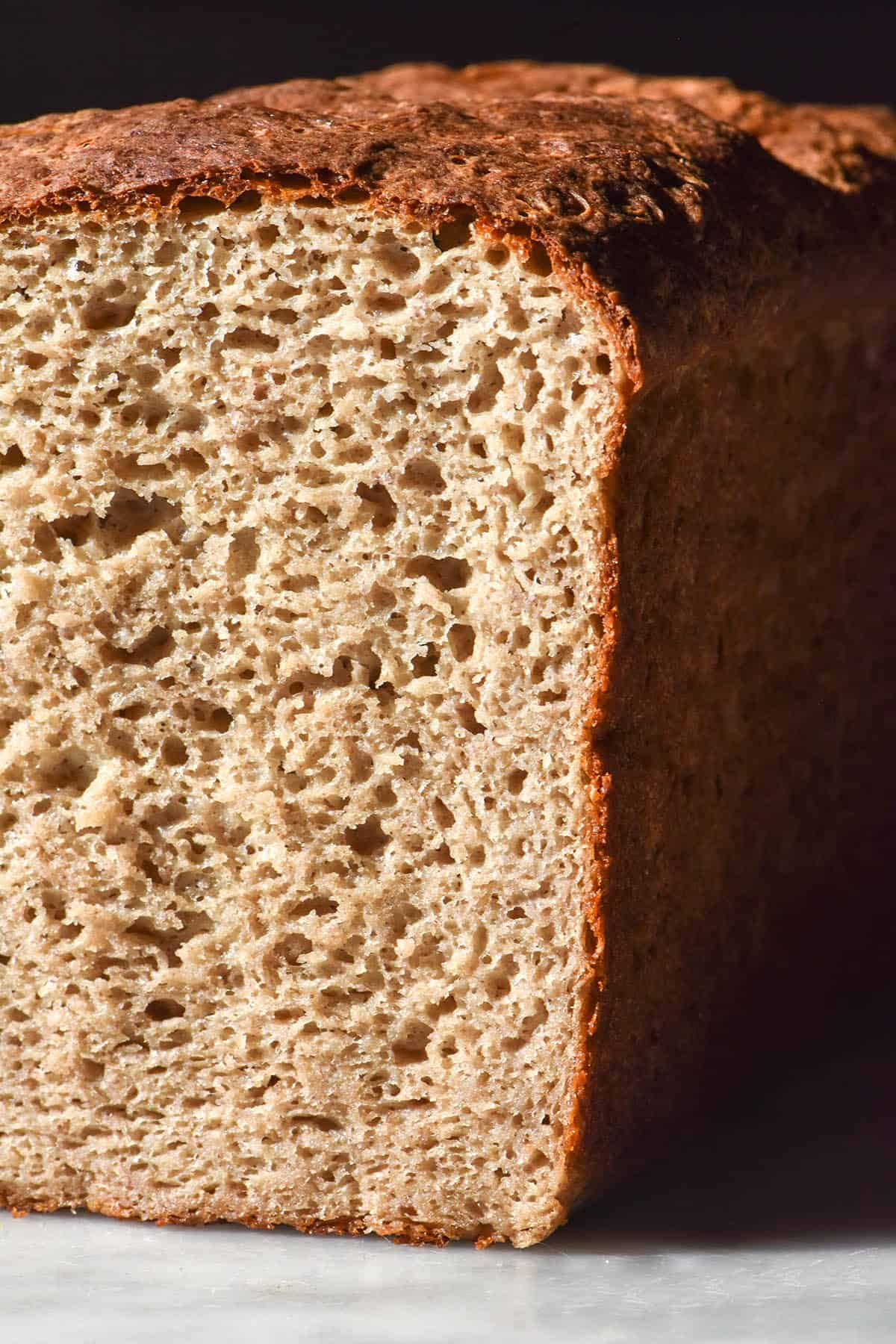 A side on image of a loaf of high protein buckwheat bread on a white marble table in contrasting sunlight. The loaf has been sliced to reveal the inner crumb.