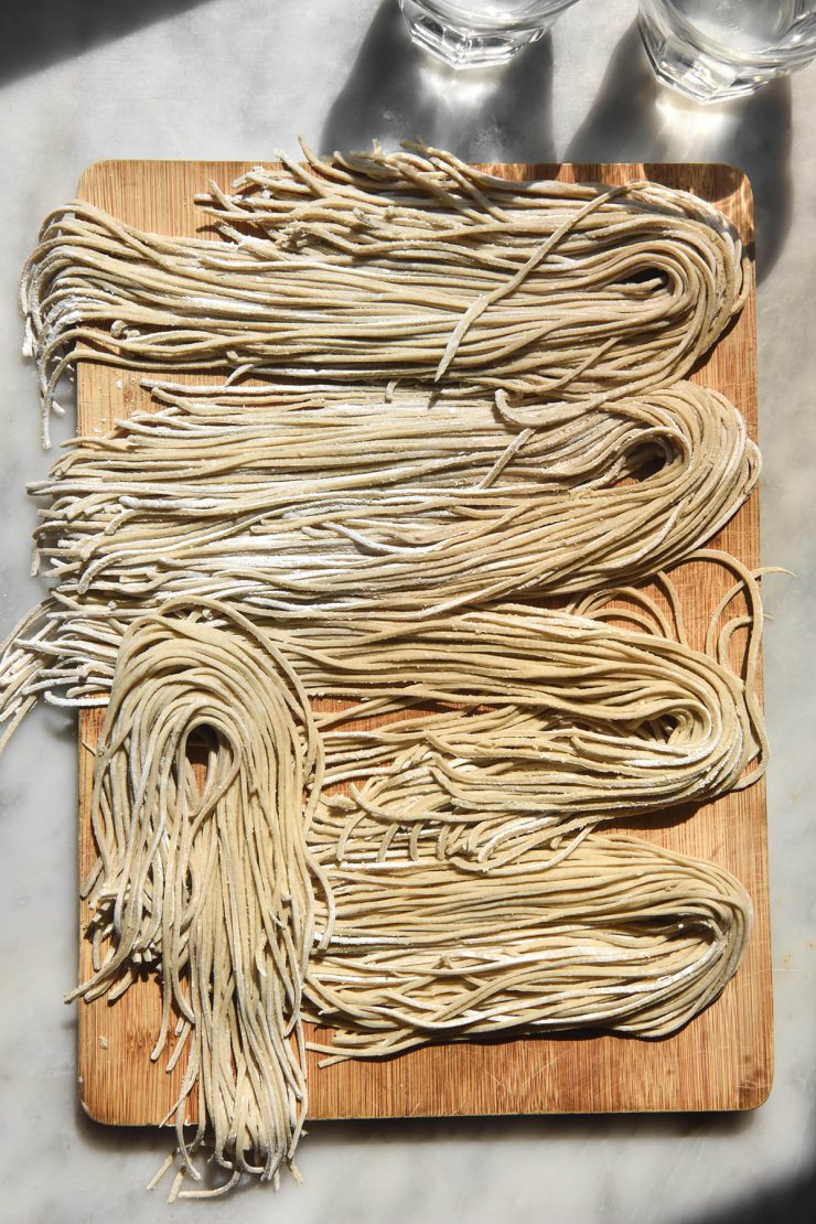 An aerial image of gluten free soba noodles arranged in groups on a wooden board atop a white marble table. The table is in contrasting bright sunlight and two glasses of water in the top of the image create a shadow and light pattern