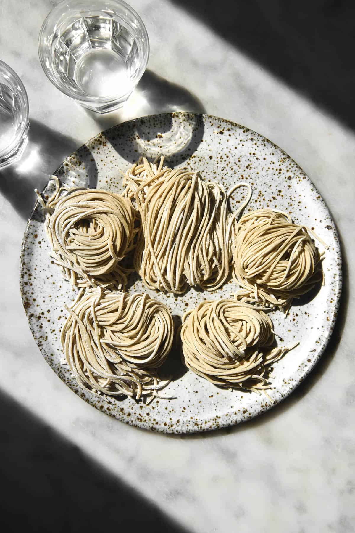 An aerial image of nests of gluten free buckwheat soba noodles on a white speckled ceramic plate atop a white marble table in contrasting sunlight. 