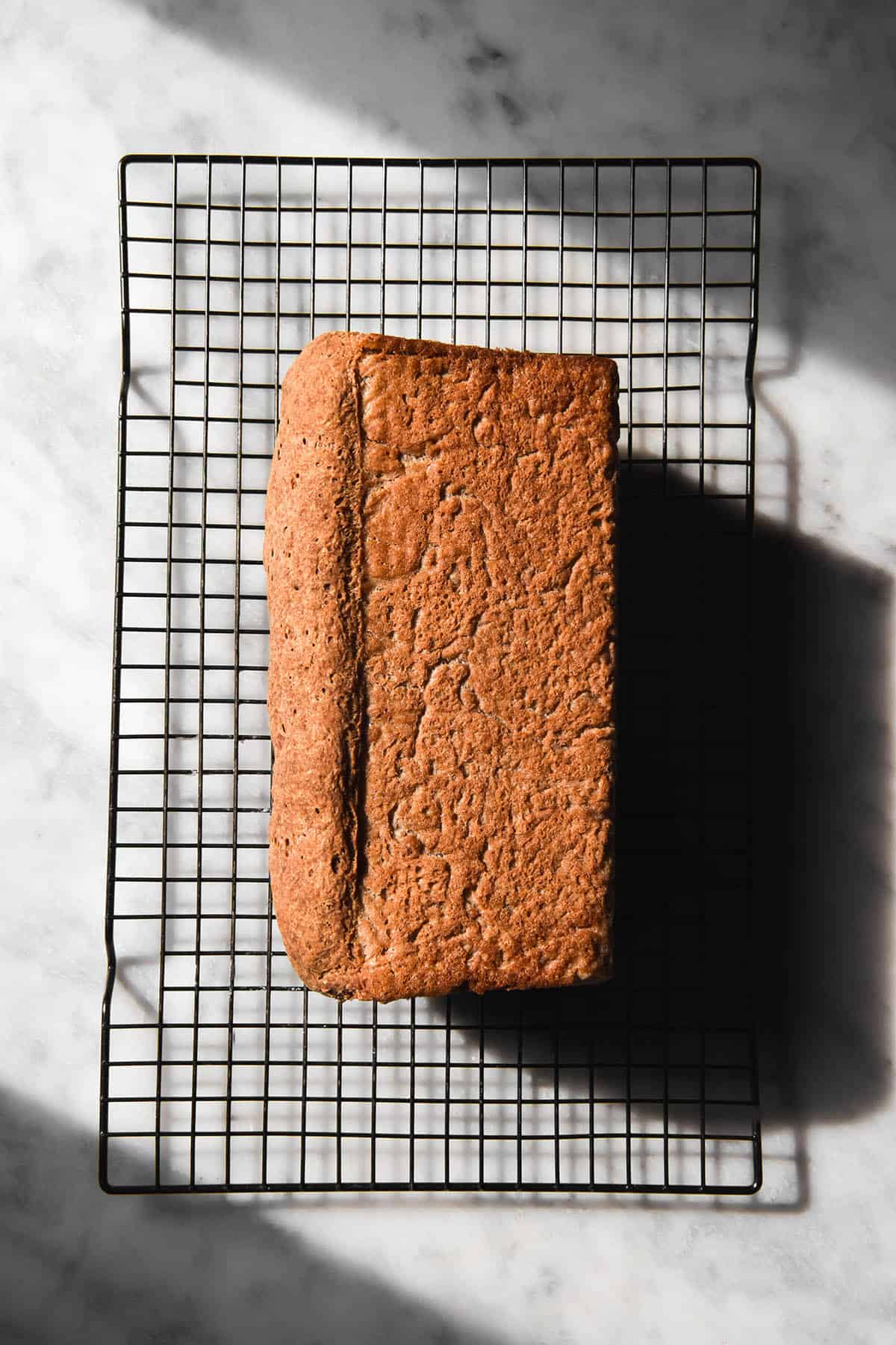An aerial image of a loaf of gluten free vegan high protein bread on a cooling rack atop a white marble table in contrasting sunlight