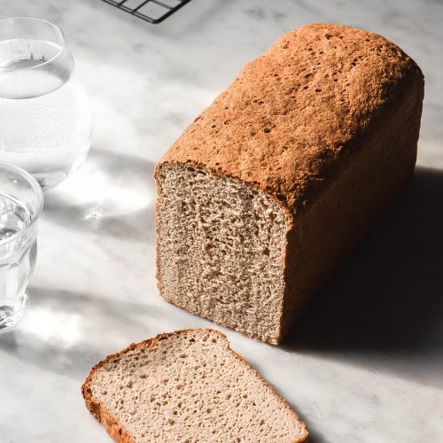 A side on image of a loaf of gluten free vegan high protein bread on a white marble table in contrasting sunlight. The bread has been sliced revealing the soft crumb. Two glasses of water sit to the left of the loaf while a cooling rack sits in the background