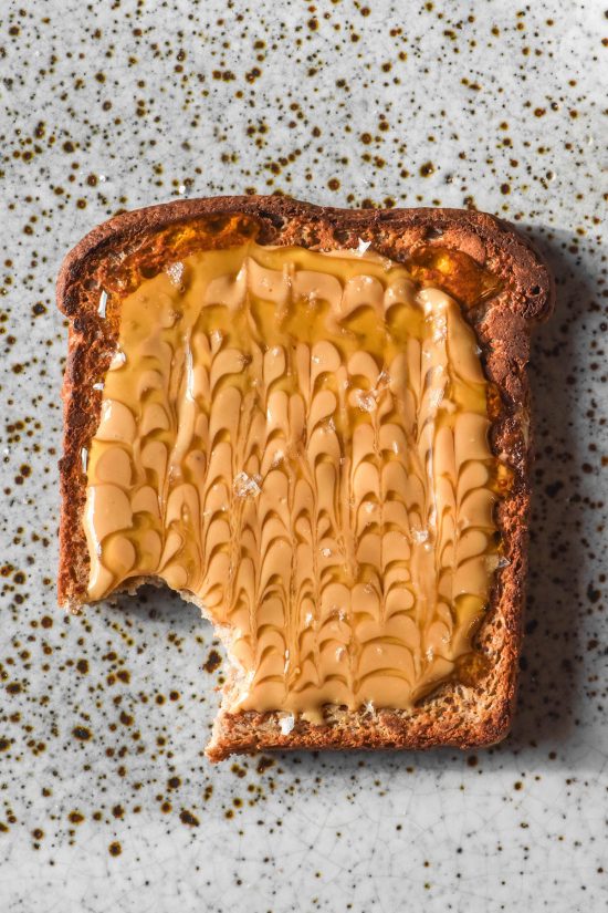 An aerial image of a slice of gluten free vegan high protein toast topped with peanut butter swirled with rice malt syrup. The toast sits atop a white speckled ceramic plate and a bite has been taken from the lower left half of the bread