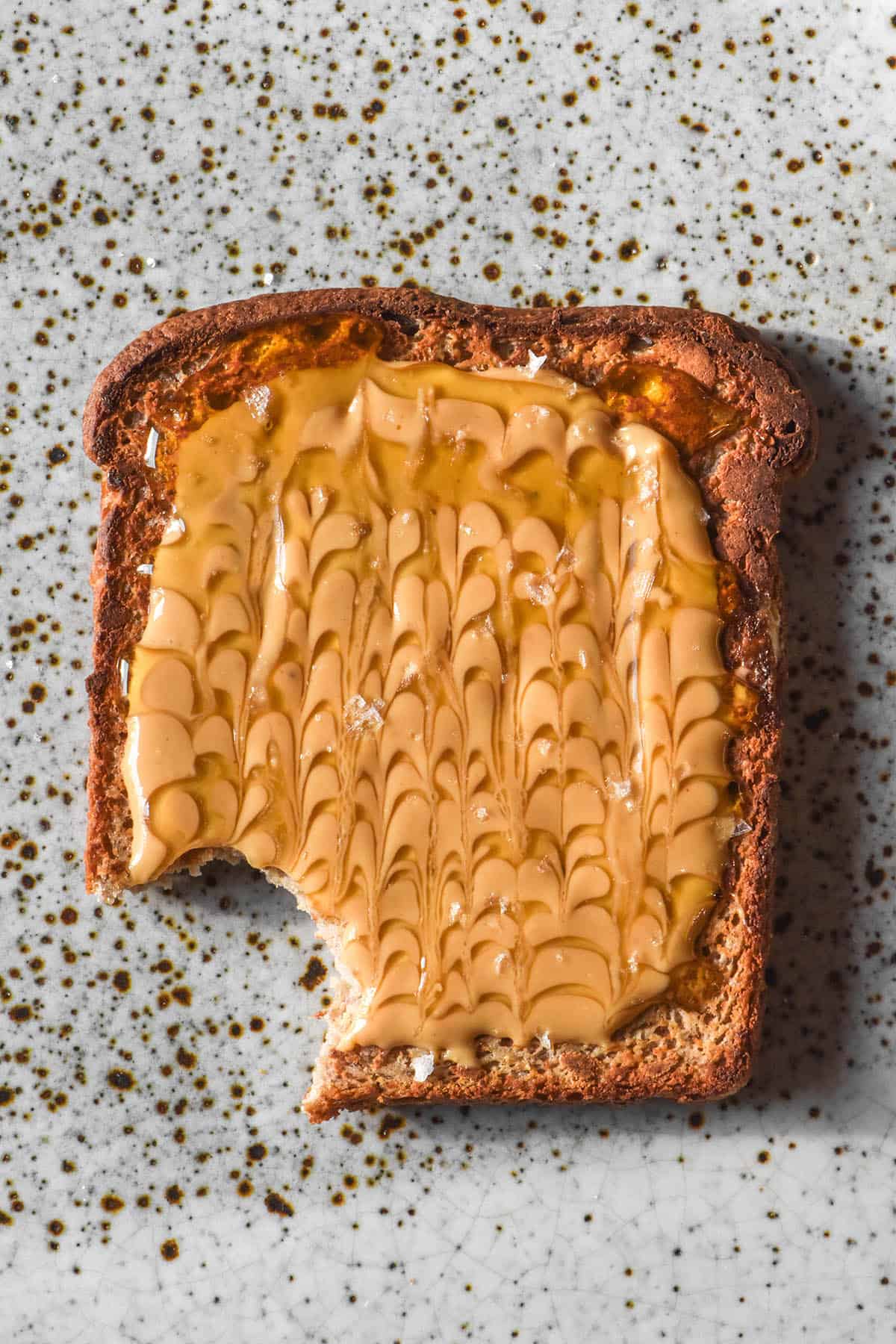 An aerial image of a slice of gluten free vegan high protein toast topped with peanut butter swirled with rice malt syrup. The toast sits atop a white speckled ceramic plate and a bite has been taken from the lower left half of the bread