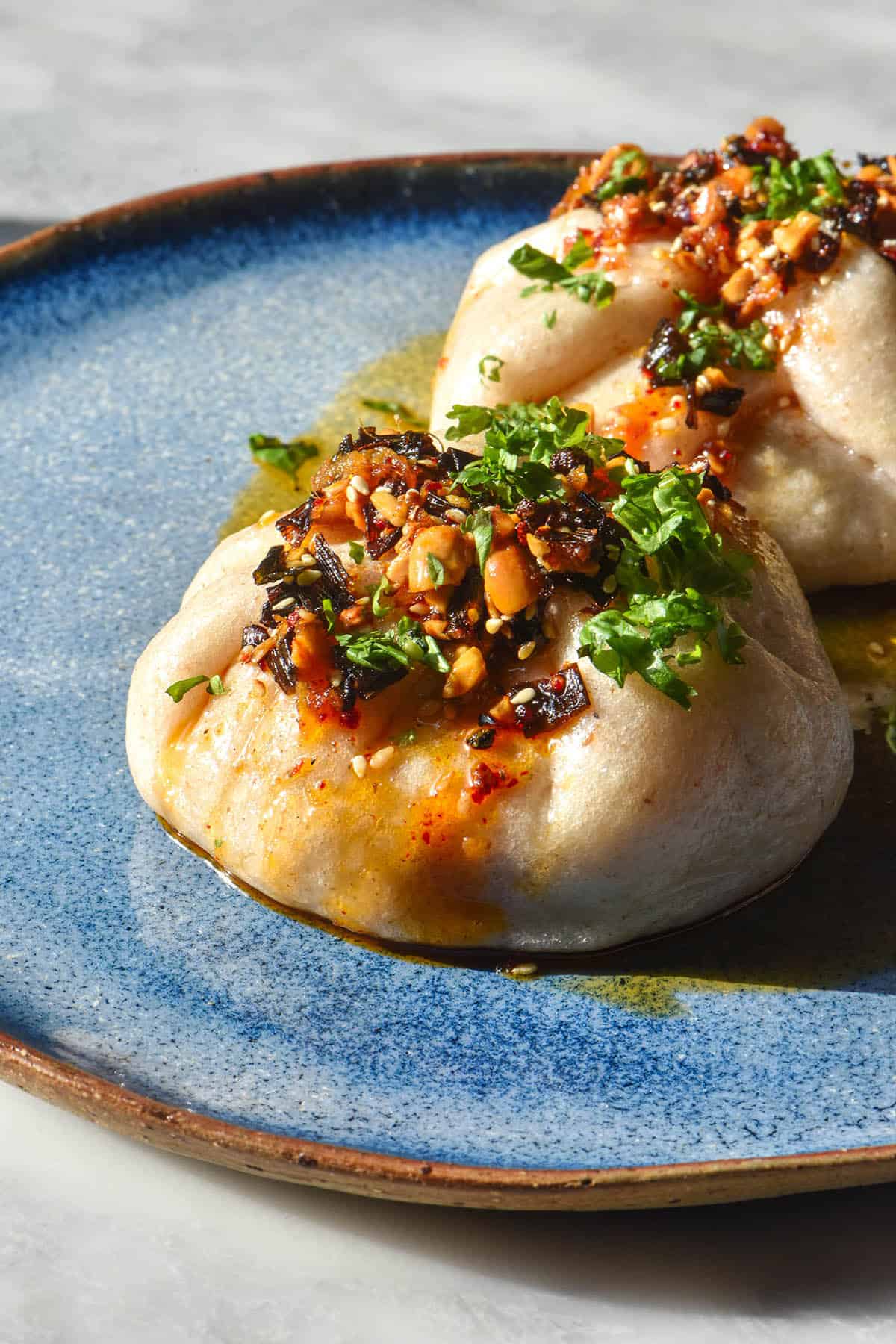 A side on image of two gluten free steamed buns topped with chilli crisp and chopped coriander. The buns sit on a bright blue ceramic plate atop a white marble table.