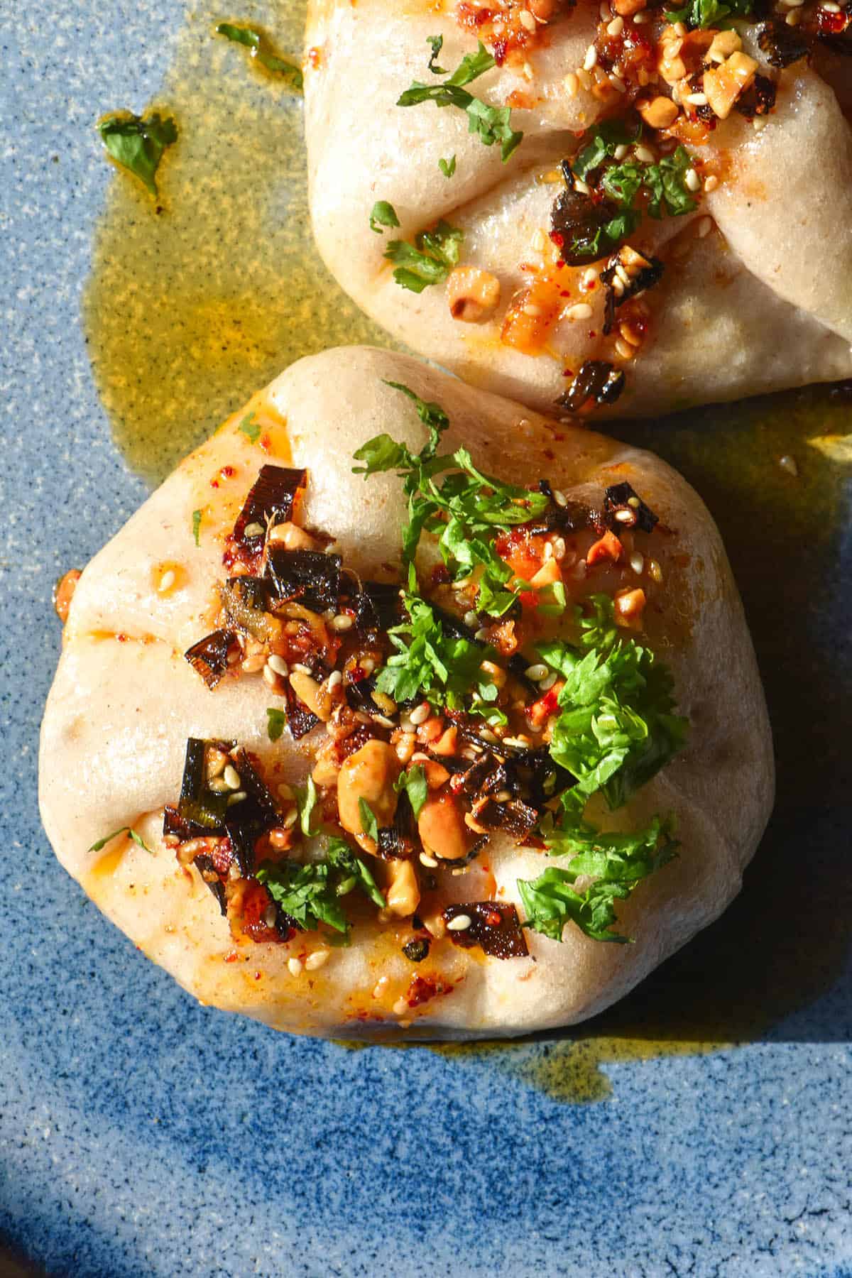 An aerial image of gluten free vegetable buns topped with chilli crisp and chopped coriander. The buns sit atop a bright blue ceramic plate in bright sunlight.