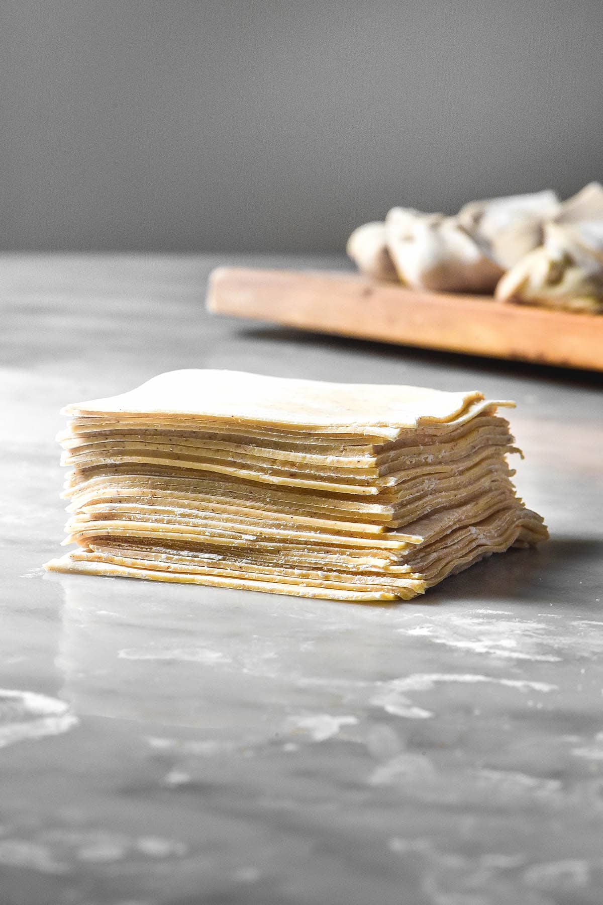An image of a stack of gluten free wonton wrappers on a white marble table against a white backdrop. A board topped with filled wontons sits in the background.