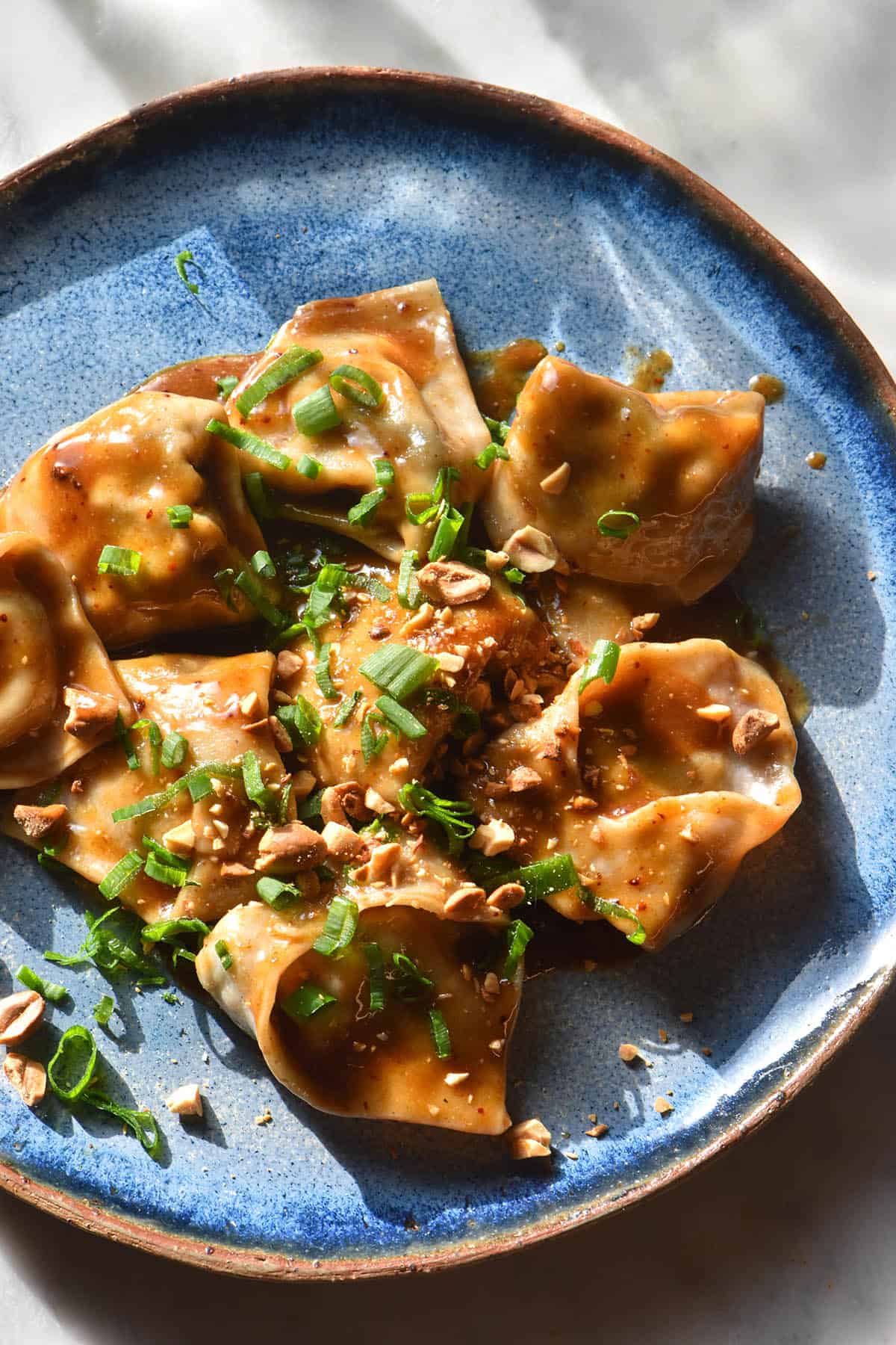 A brightly lit aerial image of gluten free wontons in a peanut butter sauce on a bright blue ceramic plate. The plate sits on a white marble table in contrasting sunlight.