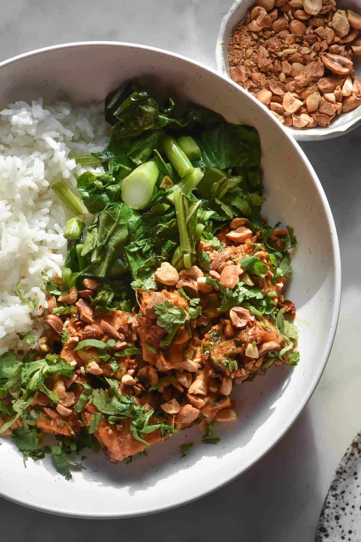 An aerial close up image of a bowl of peanut tofu with white rice and stir fried Asian greens. A bowl of toasted chopped peanuts sits to the top right of the image