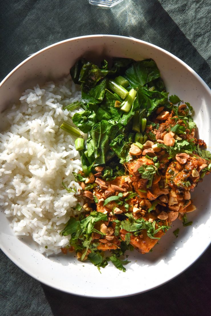 An aerial image of a white bowl filled with low FODMAP peanut tofu, white rice and Asian greens. The bowl sits atop a dark green linen tablecloth.