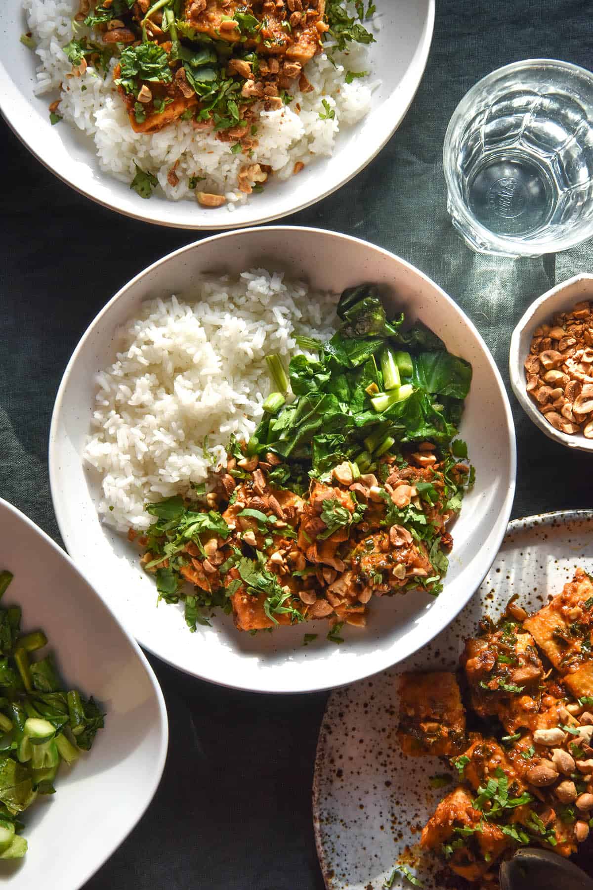 An aerial image of bowls and plates topped with low FODMAP peanut tofu atop a dark green linen tablecloth. The tofu is served with white rice and Asian greens and topped with chopped peanuts and coriander.