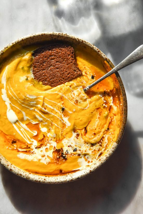 An aerial brightly lit image of a speckled ceramic bowl filled with low FODMAP carrot soup. The soup is topped with a swirl of low FODMAP chilli crisp and cream and a slice of toast. The bowl sits on a white marble table.