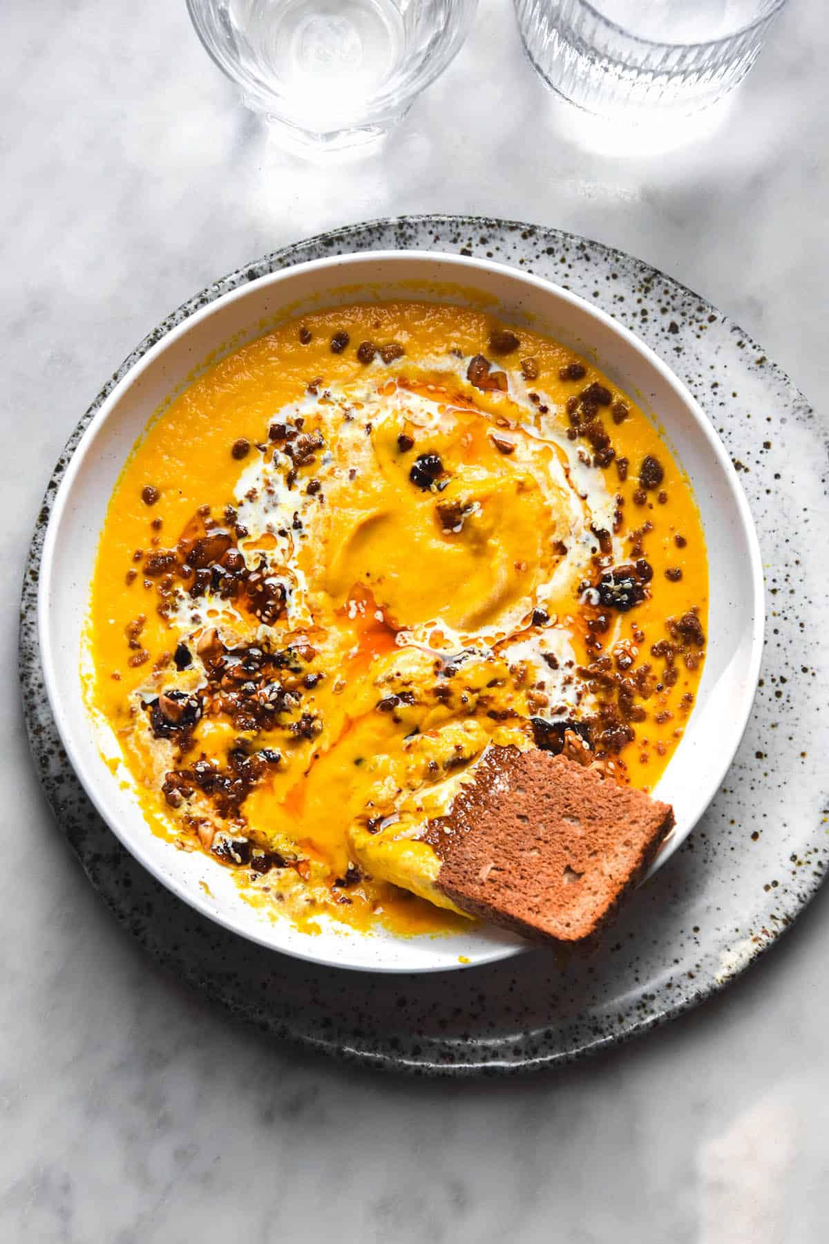 An aerial brightly lit image of a white bowl filled with low FODMAP carrot soup. The soup is topped with a swirl of low FODMAP chilli crisp, tofu crumbles and cream and a slice of toast. The bowl sits on a white speckled ceramic plate atop a white marble table.