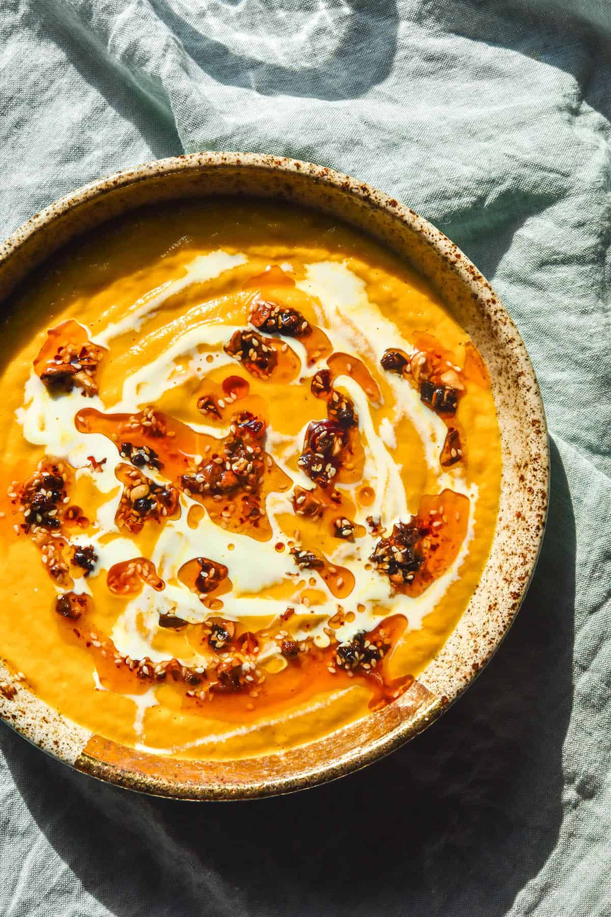 An aerial image of a speckled ceramic bowl filled with low FODMAP carrot soup. The carrot soup is topped with chilli crisp, cream and tofu crumbles and sits atop a pale green linen tablecloth.