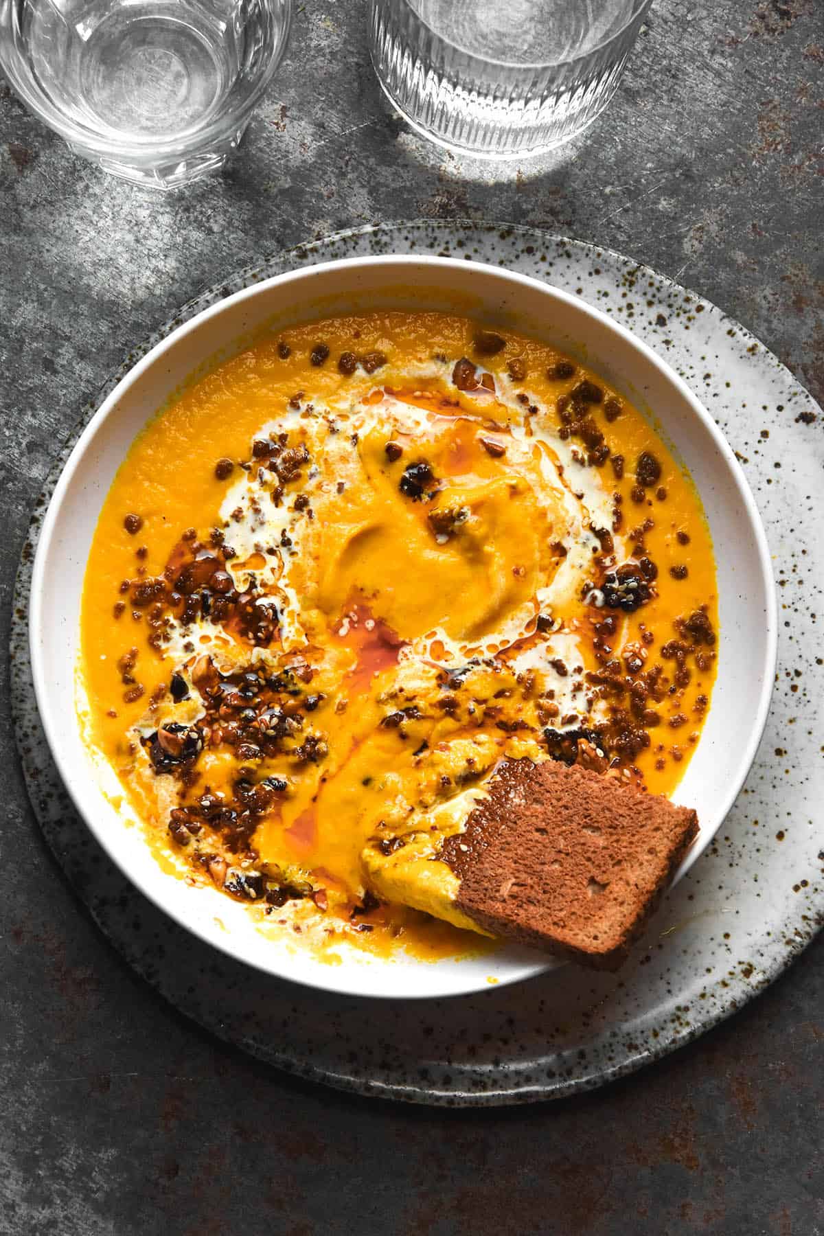 An aerial image of a white bowl filled with low FODMAP carrot soup topped with chilli crisp and cream and served with toast. The bowl sits on a white speckled ceramic plate atop a dark steel backdrop.