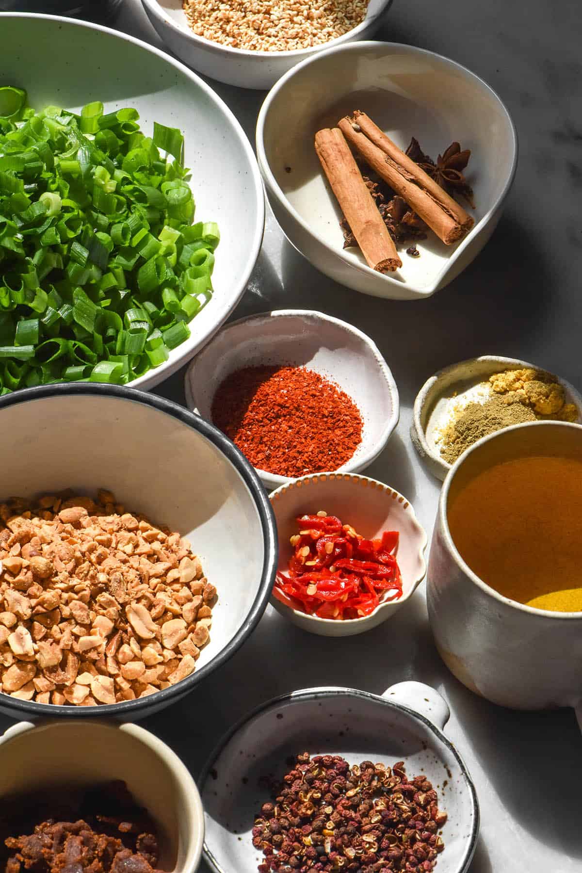 A close up image of the ingredients used to make chilli crisp in small white bowls atop a white marble table in contrasting sunlight.