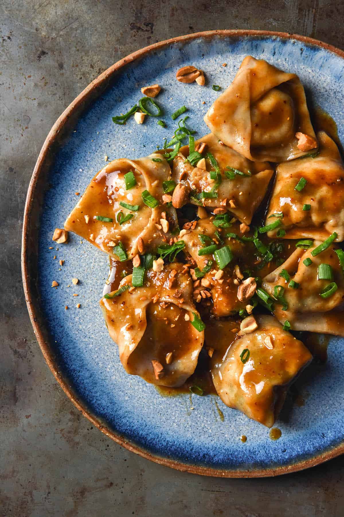 An aerial close up image of gluten free wontons topped with low FODMAP dumpling dipping sauce on a bright blue ceramic plate atop a dark grey backdrop