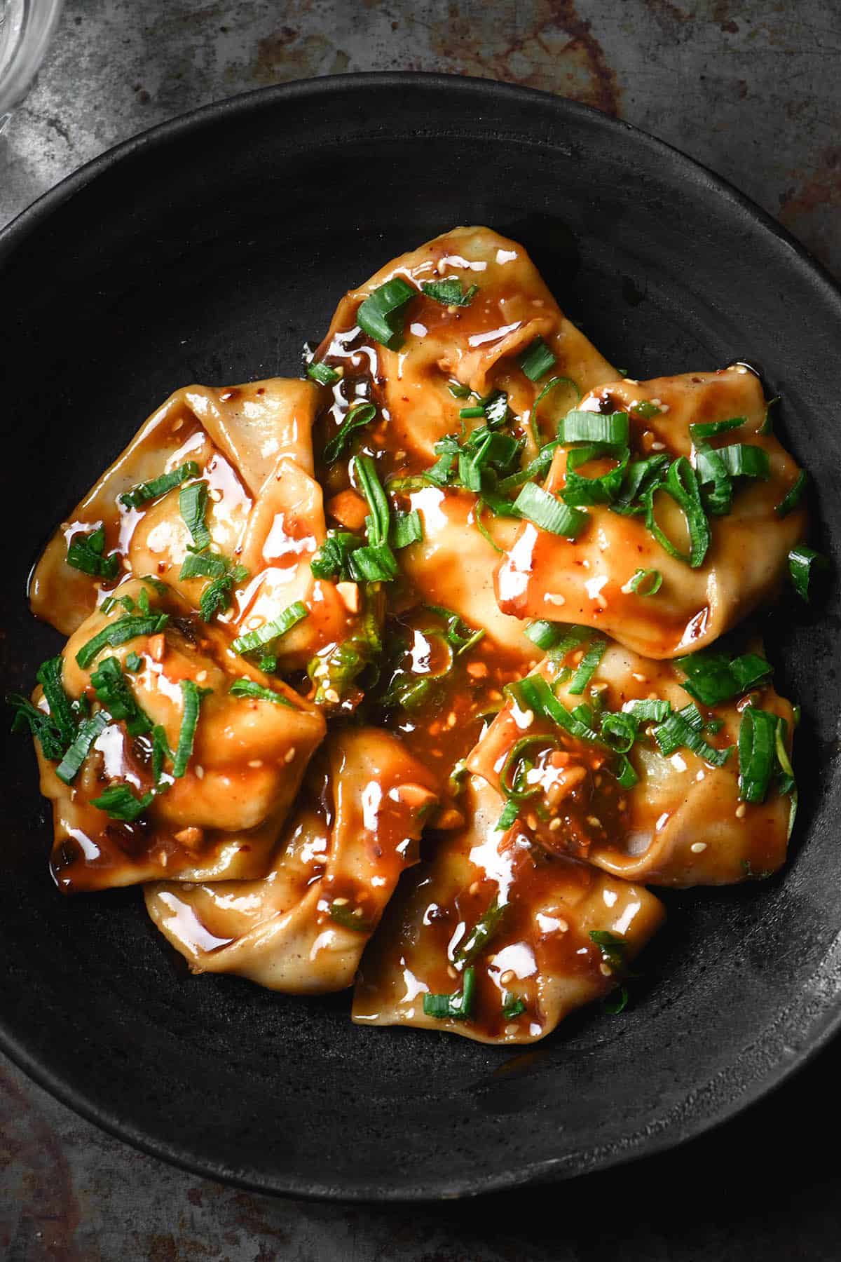 A dark and moody aerial image of gluten free wontons in a dark ceramic bowl topped with a low FODMAP dumpling sauce. The bowl sits atop a dark steel backdrop