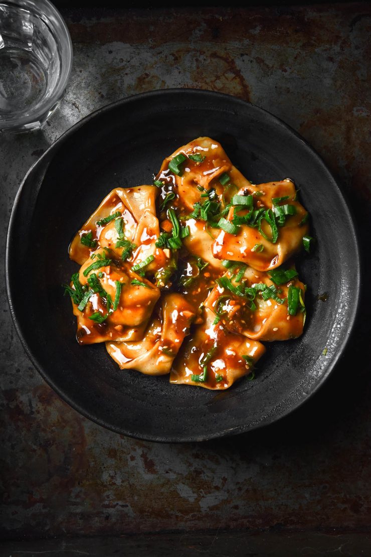 A dark and moody aerial image of gluten free wontons in a dark ceramic bowl topped with a low FODMAP dumpling sauce. The bowl sits atop a dark steel backdrop and a glass of water sits to the top left of the image