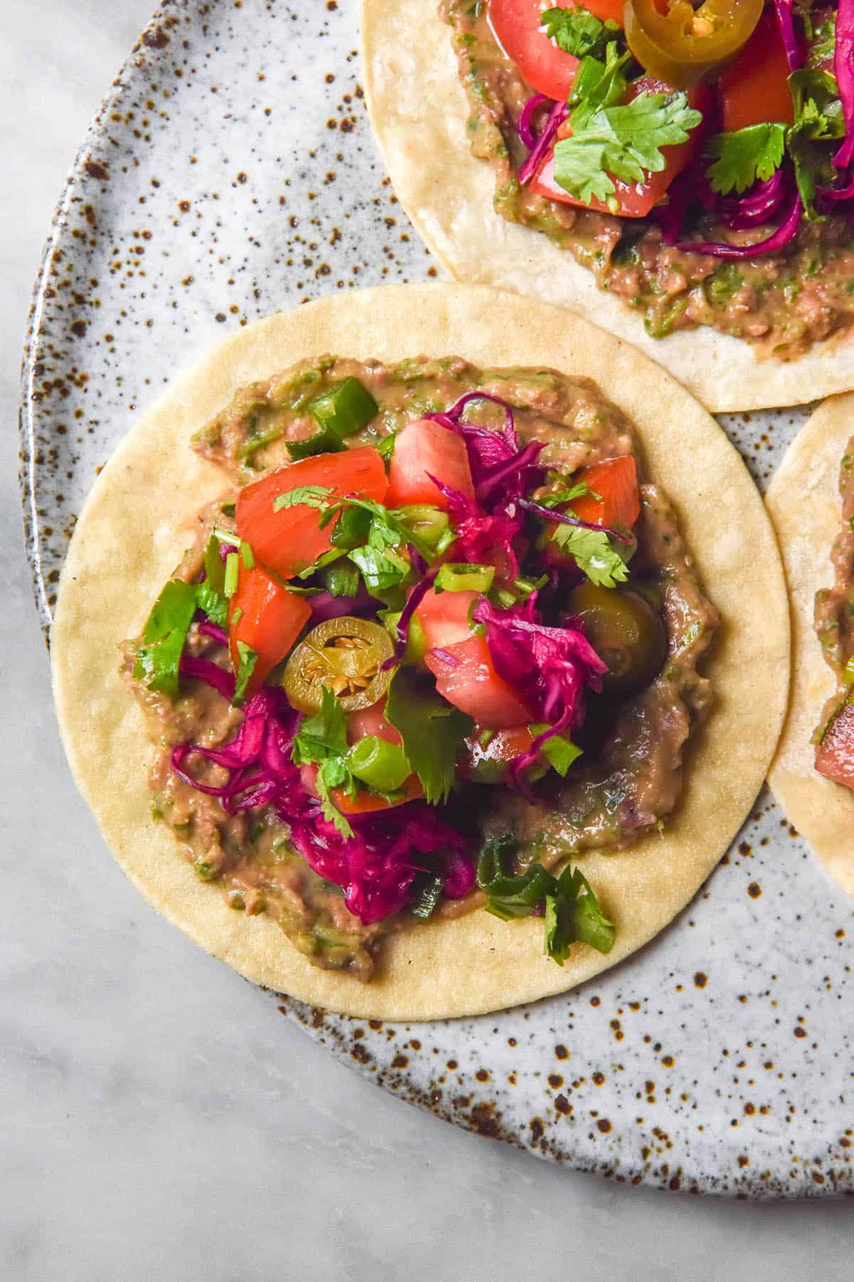 An aerial image of corn tacos on a white speckled ceramic plate topped with low FODMAP refried beans, pickled vegetables, tomato, jalapeno and coriander.