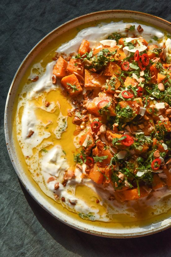 An aerial image of a white ceramic plate topped with Thai red curry roasted pumpkin on a bed of yoghurt, topped with peanuts, herbs, chilli and a lime coriander salsa. The plate sits atop a dark green linen tablecloth.